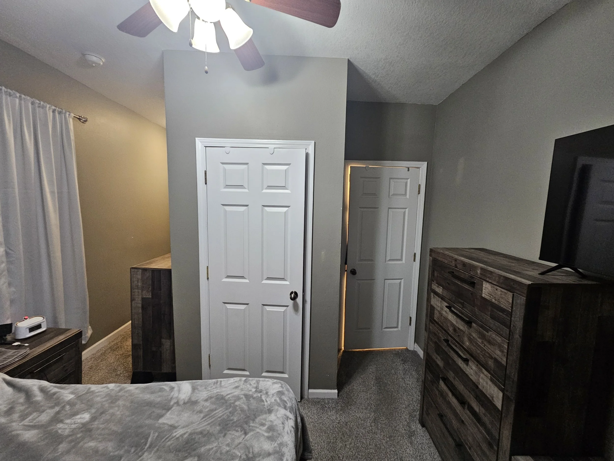 Interior view of a bedroom corner with a ceiling fan, two closed white closet doors, a wooden dresser, a small table with a device, and a window with white curtains.