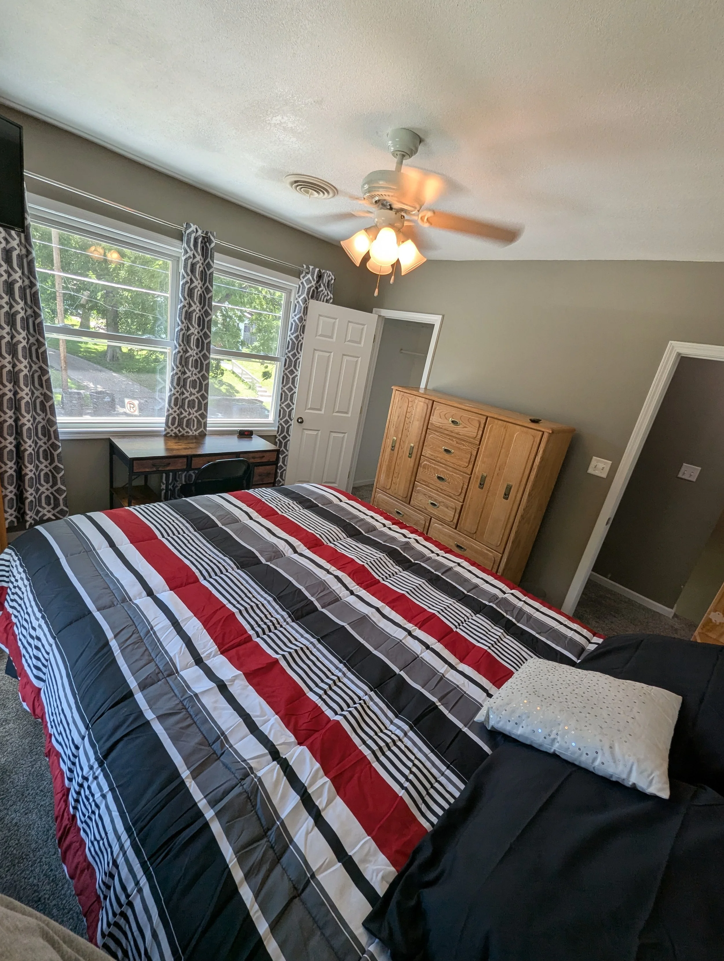 A bedroom with two large windows with grey patterned curtains, a wooden dresser, a ceiling fan with lights turned on, a bed with a multi-colored striped quilt, and a decorative pillow with sequins.