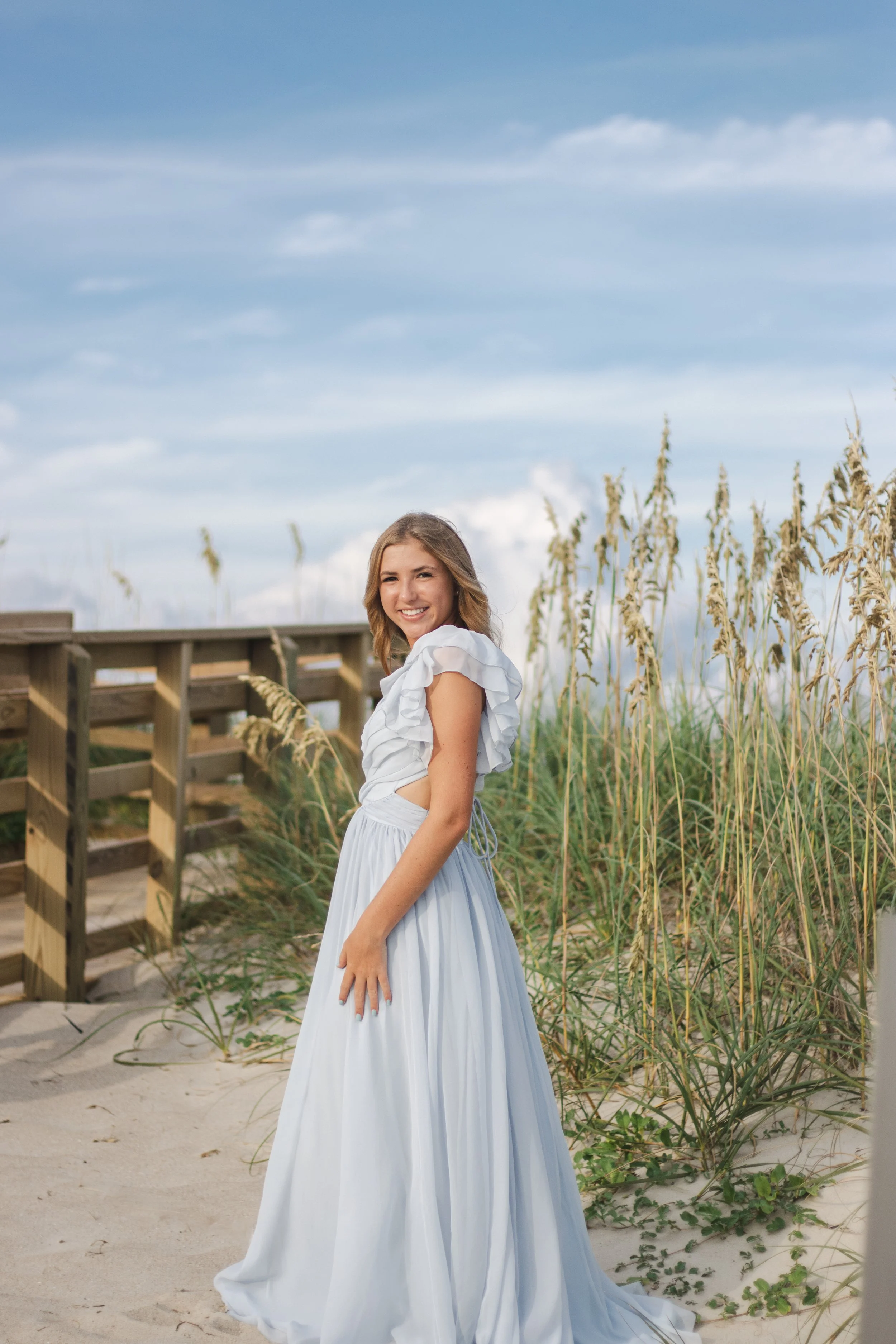 A young woman in a white dress with ruffled sleeves standing on a sandy beach near dunes and tall grasses, smiling at the camera under a partly cloudy sky.