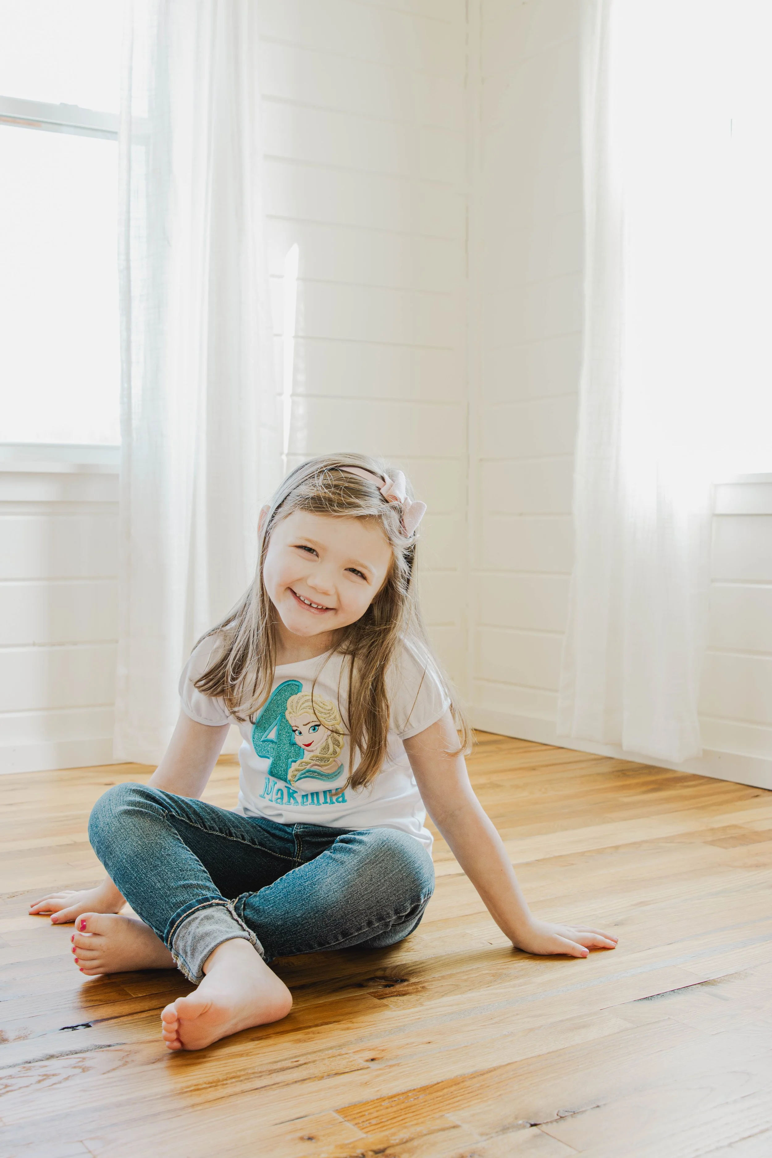 A young girl with long hair, smiling, sitting on a wooden floor in a bright room with white curtains and walls. She wears a white t-shirt with a princess graphic and the number 4, and blue jeans.
