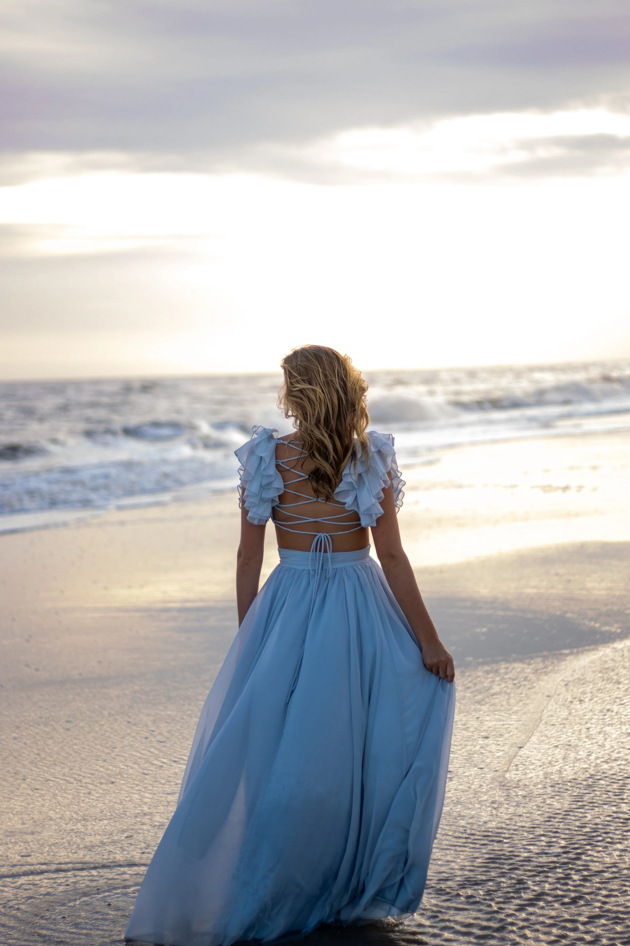 Woman in a flowing blue dress walking on the beach near the ocean at sunset.