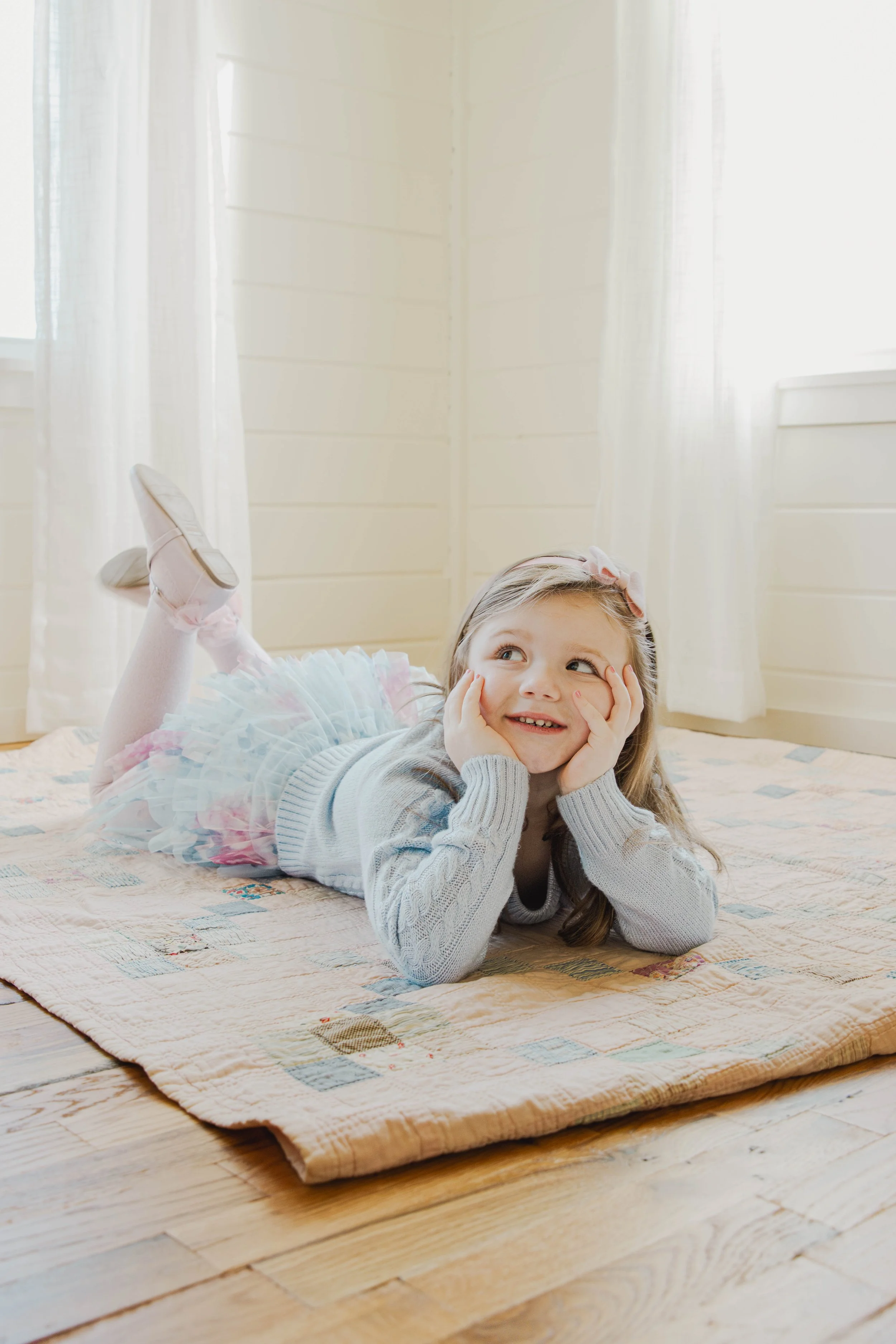 A young girl lying on her stomach on a quilted rug, smiling and resting her head on her hands, in a brightly lit room with white curtains and walls.