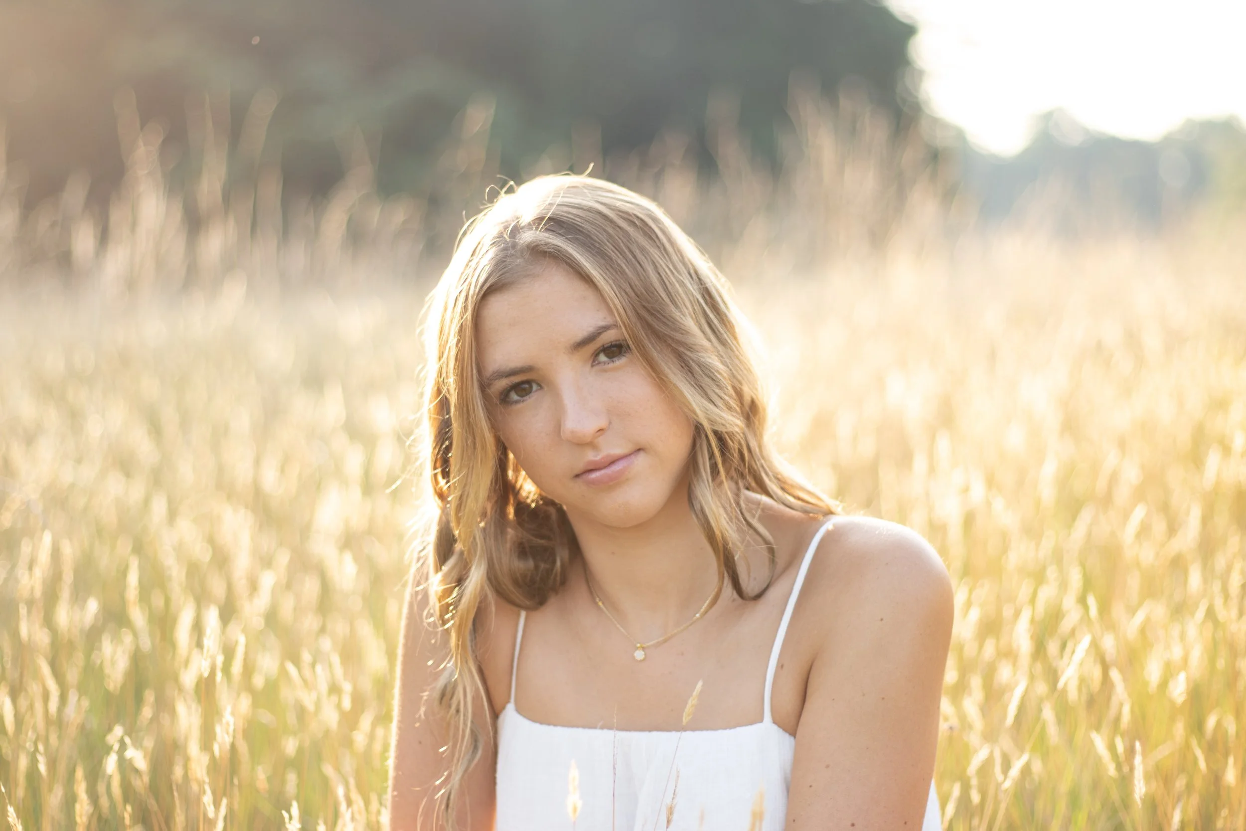 Young woman with blonde hair in a white dress sitting in a sunlit field of tall grass.