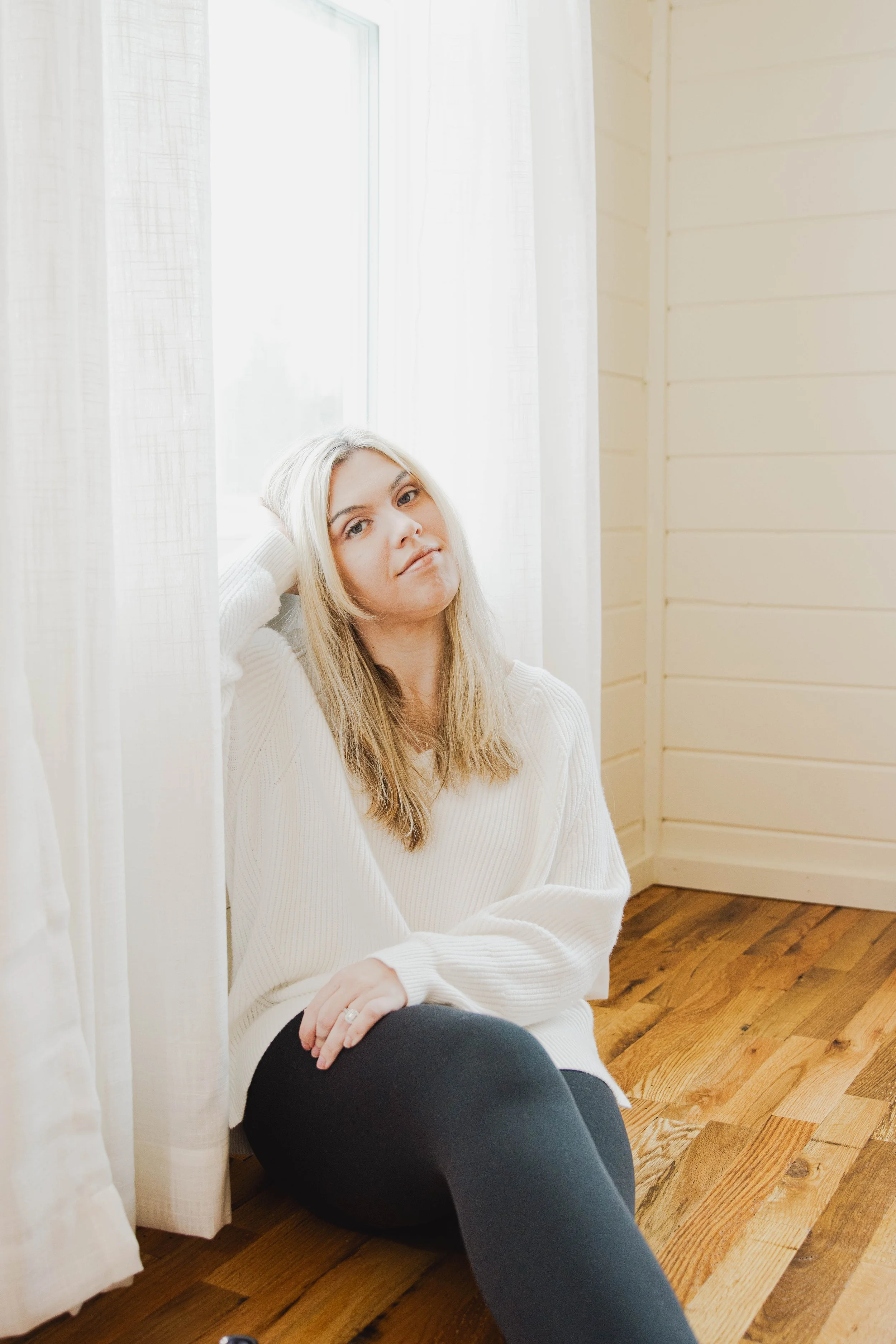 A young woman with blonde hair sitting on a wooden floor next to a window with white curtains, wearing a white sweater and black pants.