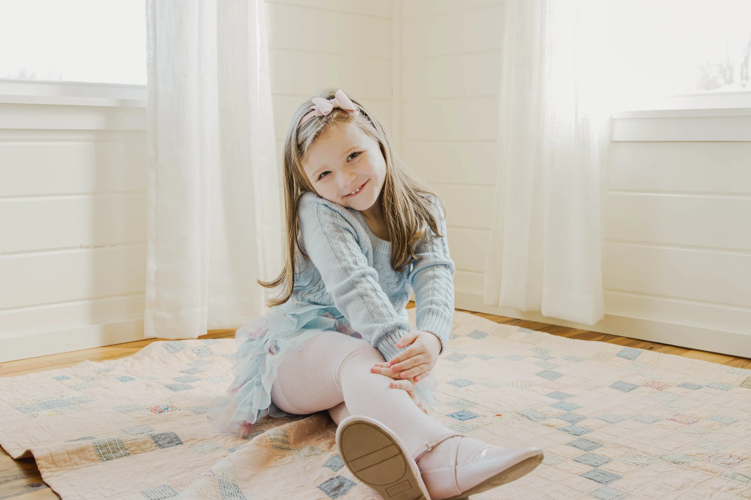 A smiling young girl with light brown hair wearing a gray sweater, pink tights, a pastel-colored tutu, and ballet shoes, sitting on a patterned rug in a bright room with white wooden walls and curtains.