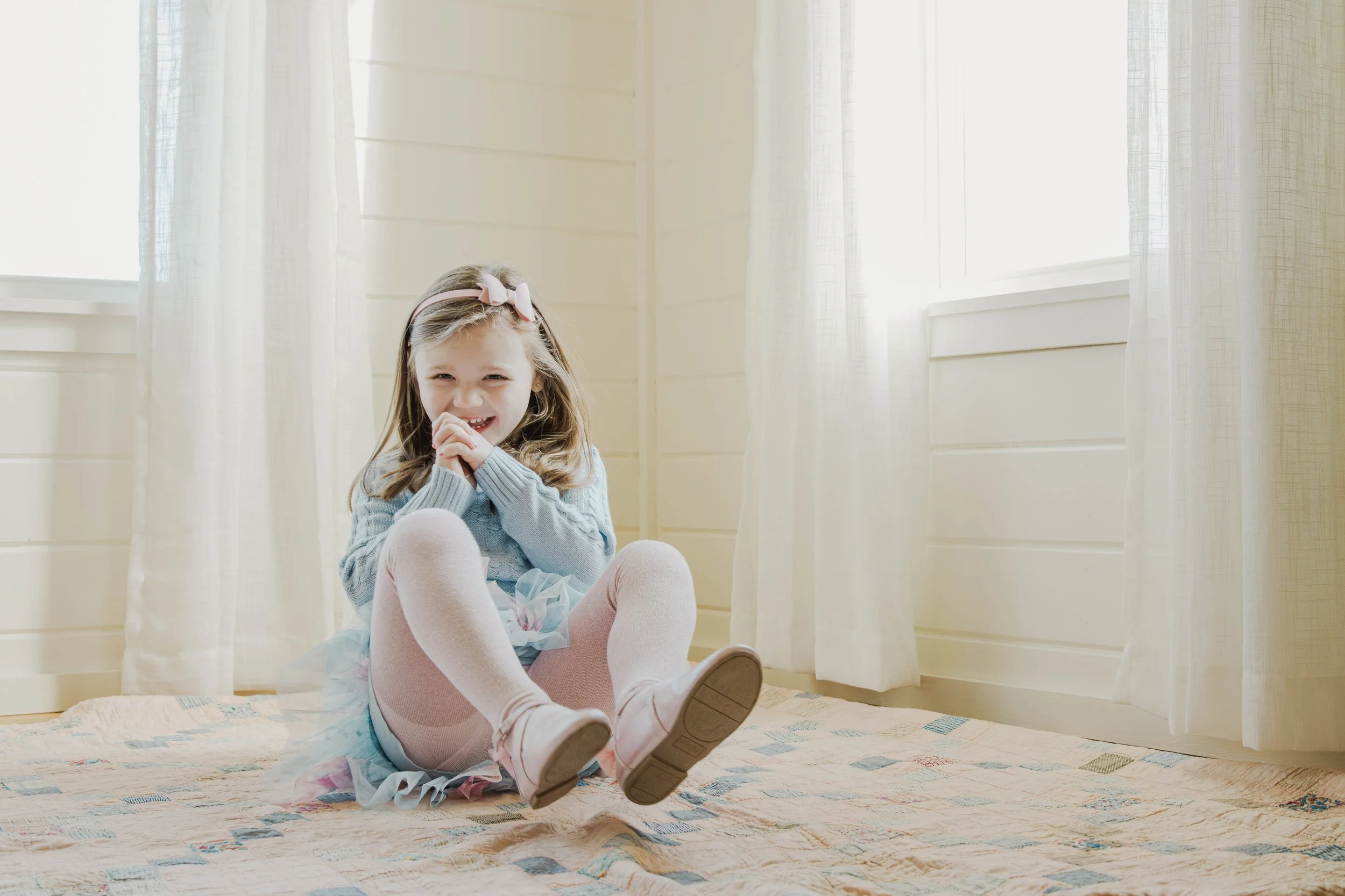 A young girl laughing while sitting on the floor in a sunlit room, wearing a sweater, tutu, tights, and shoes.