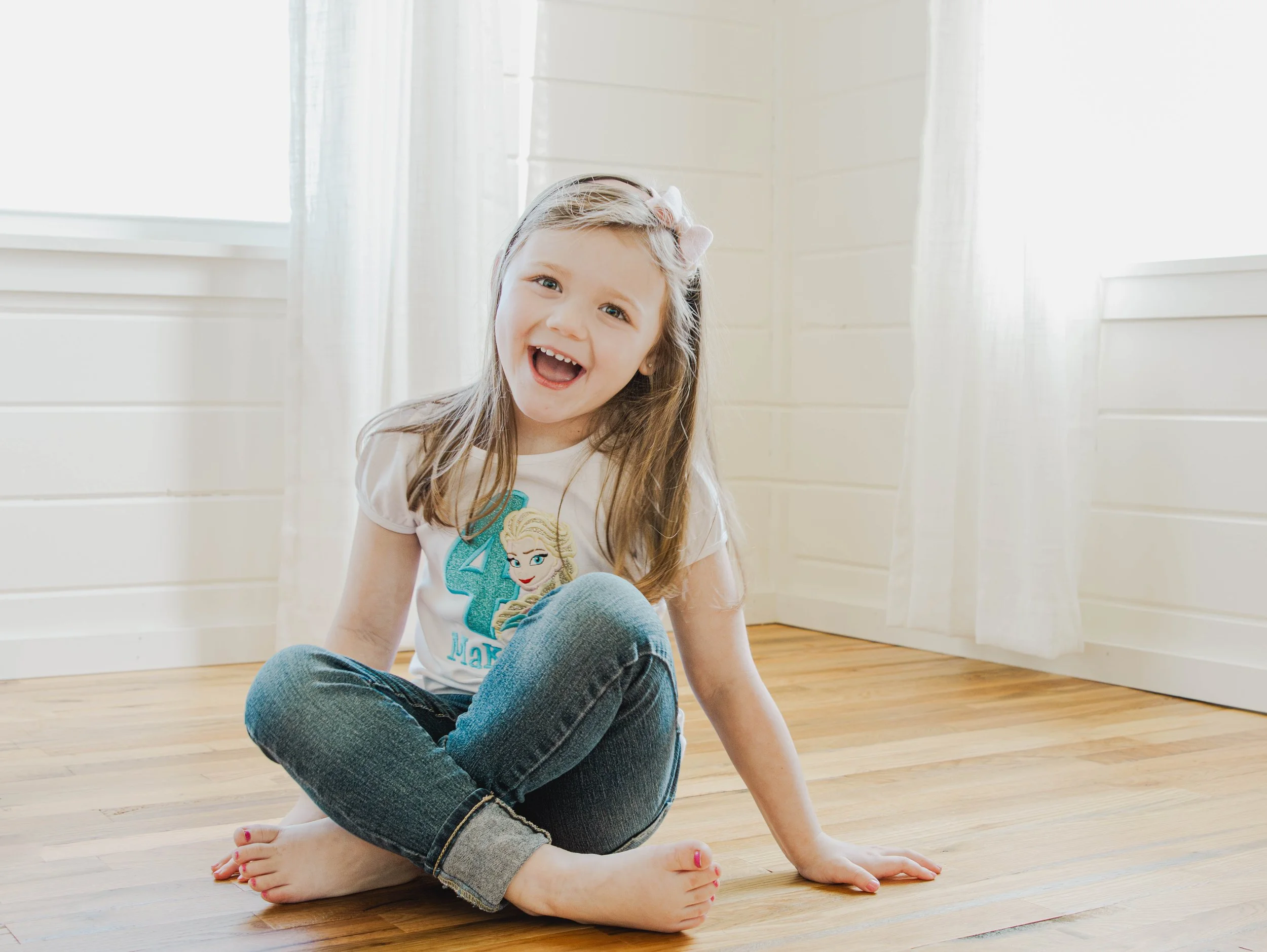 A young girl with long brown hair, smiling and sitting cross-legged on a wooden floor in a bright room with white walls and curtains, wearing a white t-shirt with a princess graphic and blue jeans.