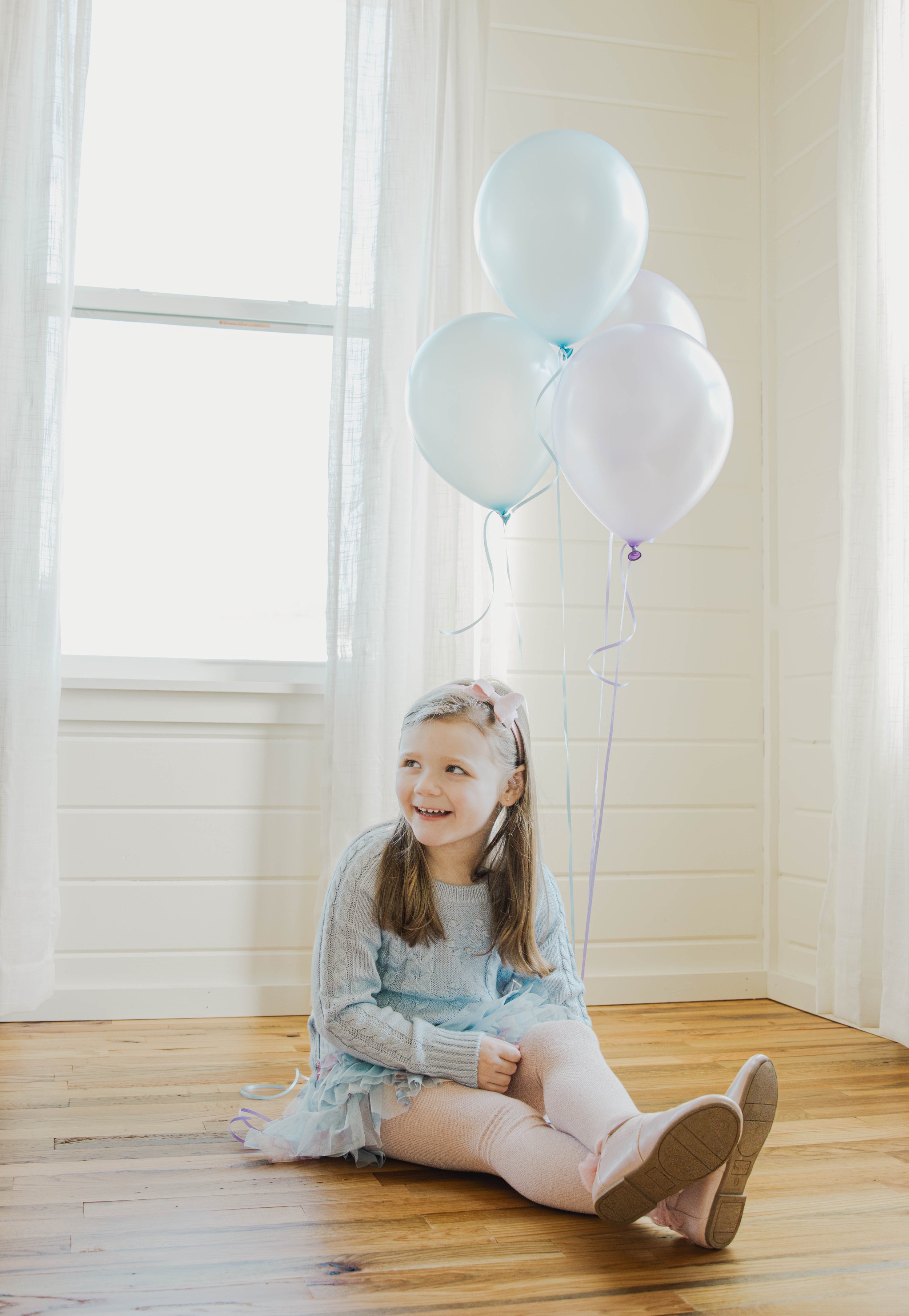 A young girl sitting on wooden floor with four pastel-colored balloons in a room with white walls and curtains, smiling.
