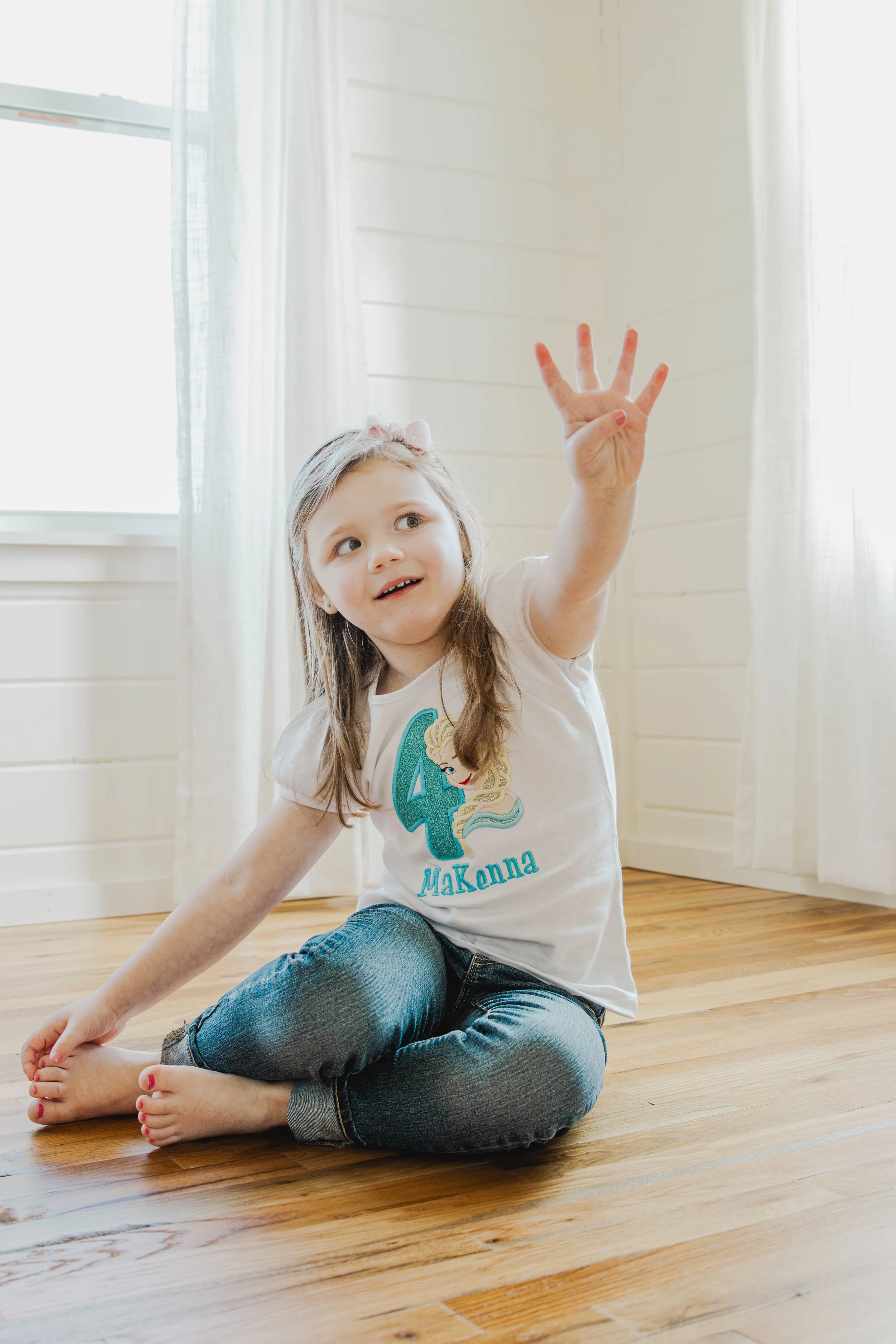 A young girl sitting cross-legged on a wooden floor, reaching up, wearing a white t-shirt with a large number 4 and a princess character, and jeans.