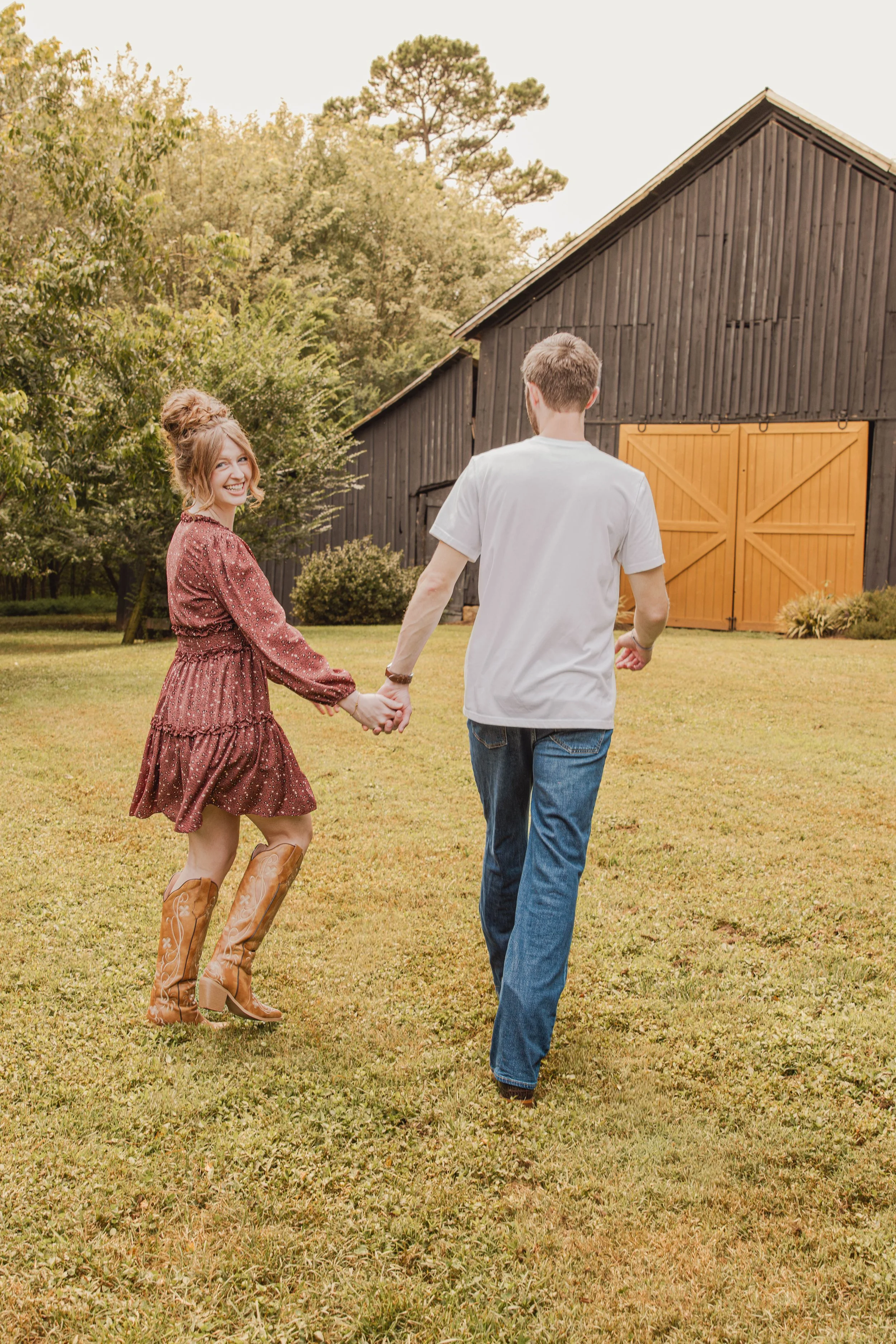 A young couple holding hands and walking on a grassy field near a black barn with orange doors, surrounded by trees.