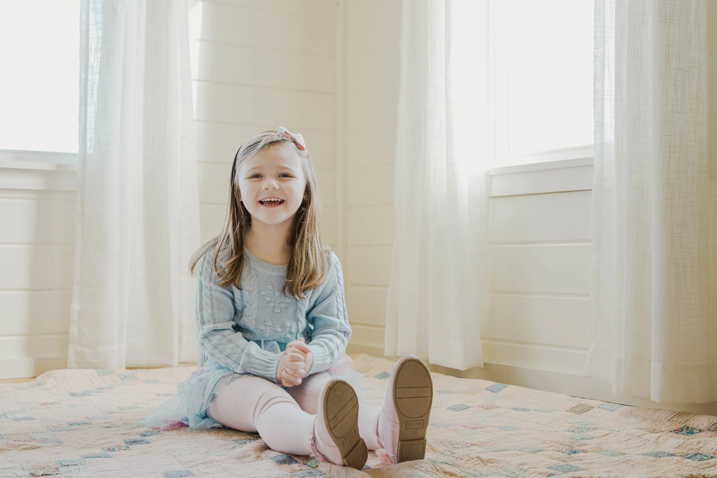 A young girl with long hair, wearing a light blue sweater and pink tights, sitting on a bed with a colorful quilt, smiling at the camera inside a bright room with white curtains and walls.