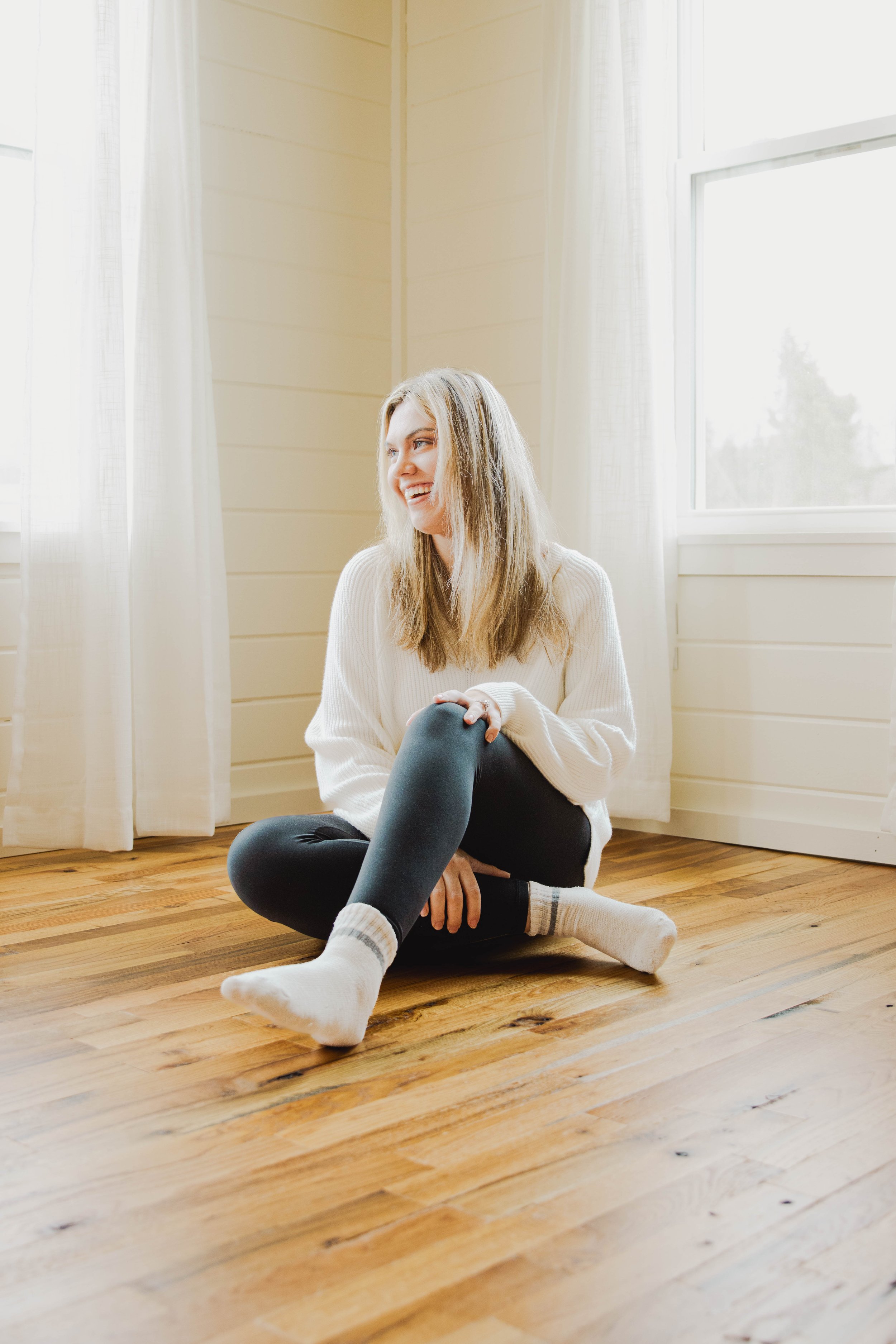 A young woman with blonde hair, wearing a white sweater, black leggings, and white socks, sitting on a wooden floor near a window, smiling and looking to the side.