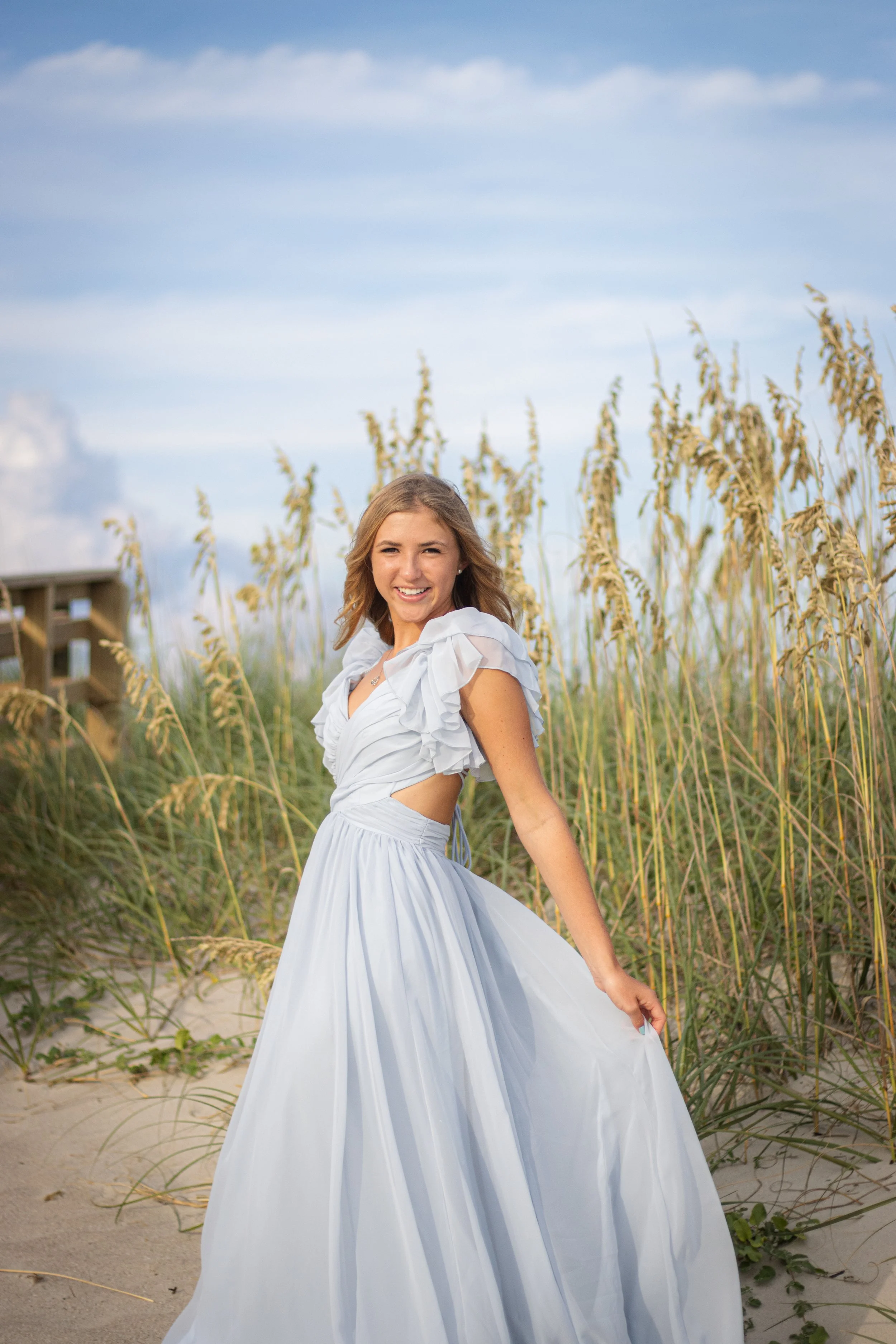 A young woman wearing a light blue dress is standing on a sandy beach with tall grass, smiling at the camera during daylight.