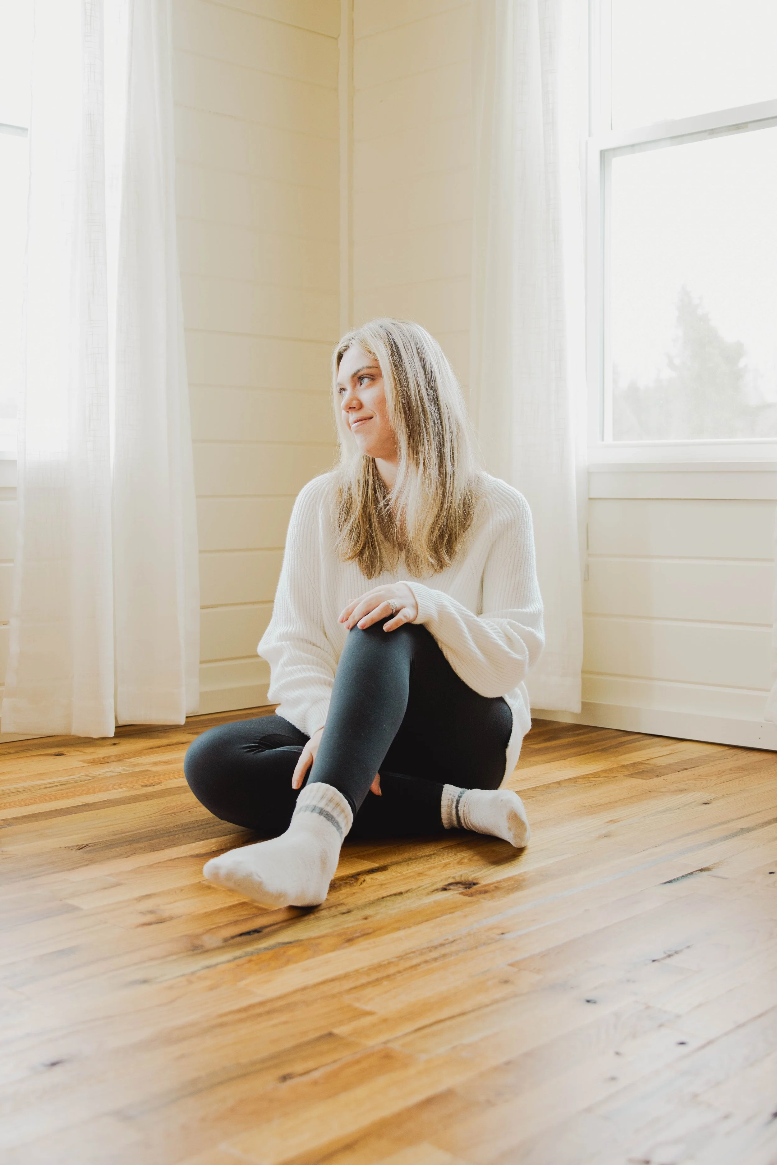 A young woman with blonde hair, wearing a white sweater, black leggings, and white socks, sitting on a wooden floor in a bright room with large windows and white curtains.