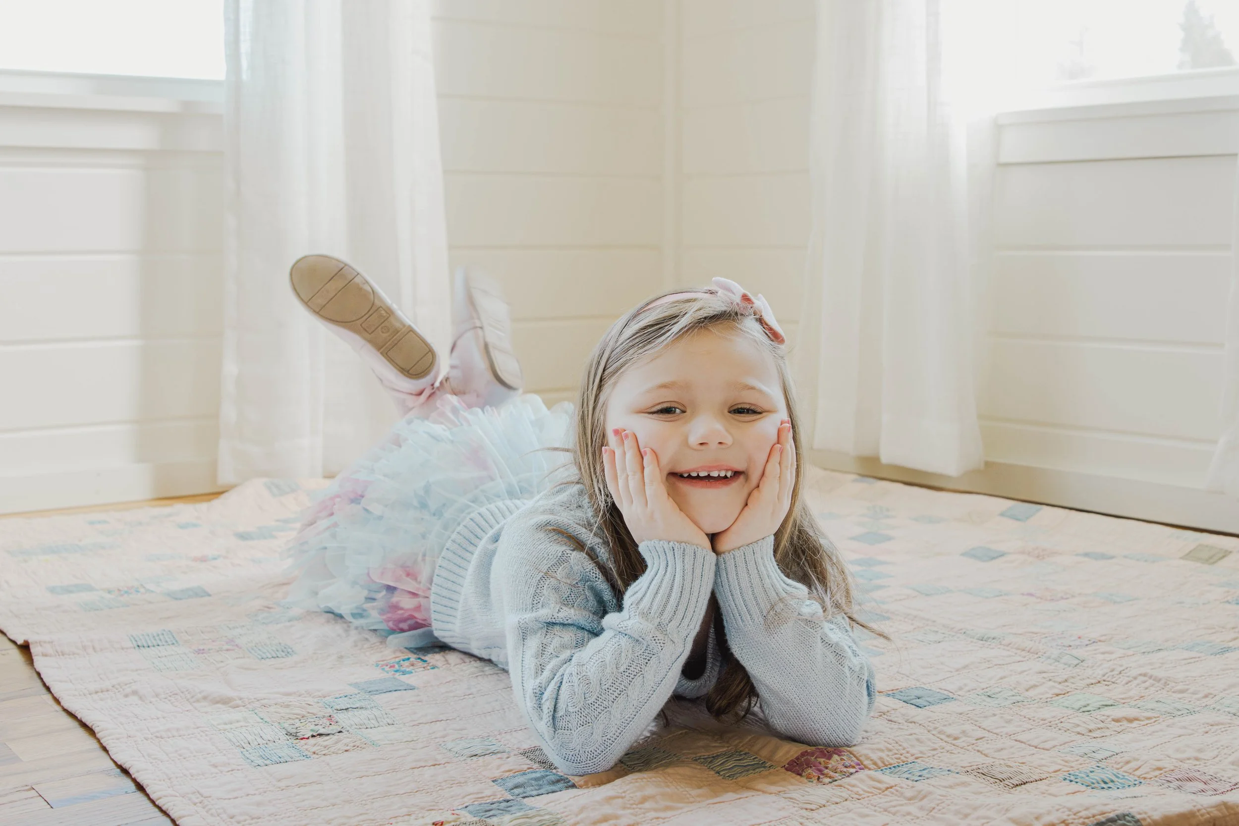 A young girl lying on her stomach on a quilted rug, smiling with her hands on her cheeks, in a bright room with white curtains.