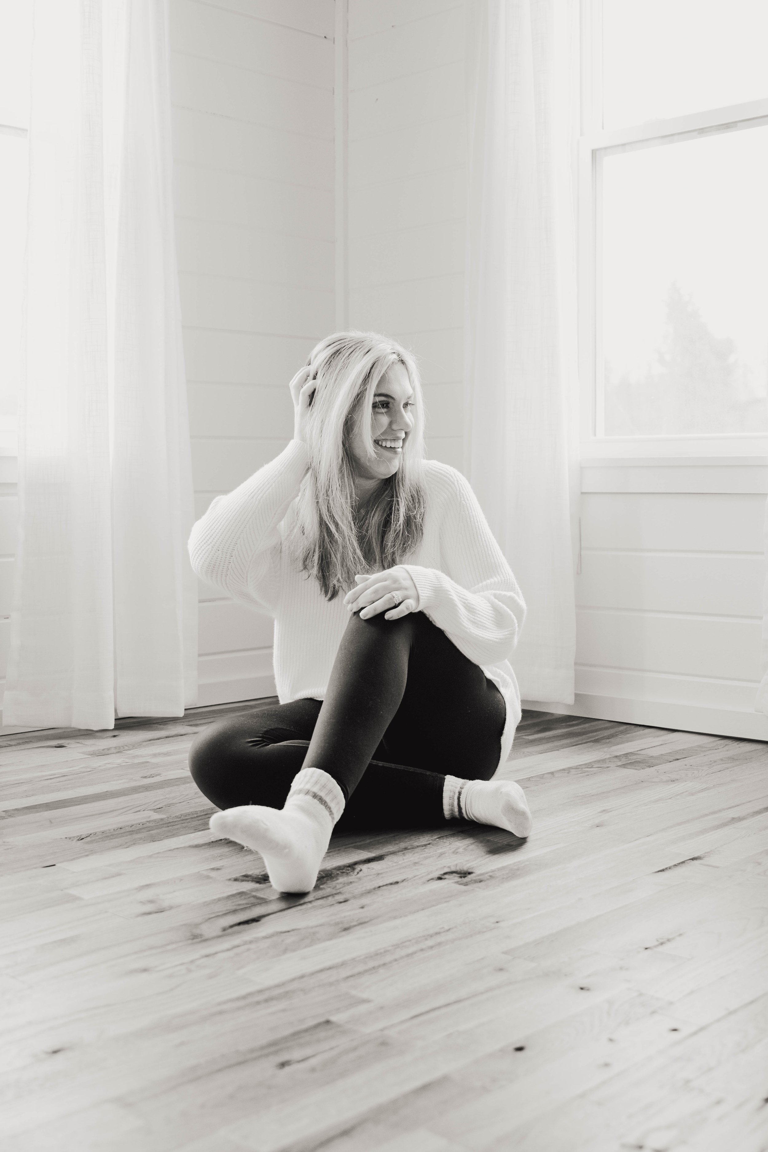 A woman sitting on a wooden floor near a window with white curtains, smiling and touching her hair.
