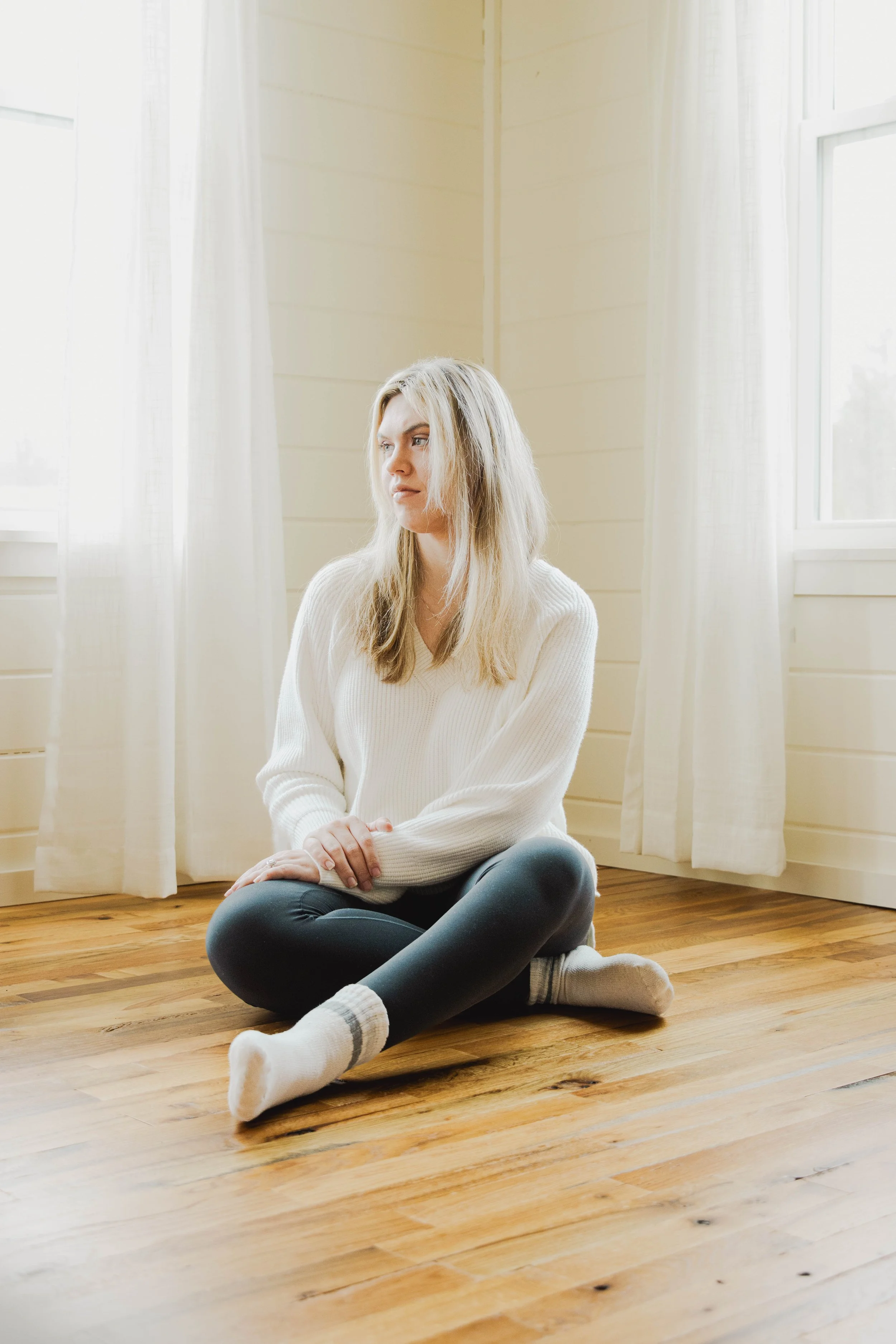 A young woman with blonde hair and a white sweater sitting on a wooden floor near a window with white curtains, looking thoughtfully into the distance.
