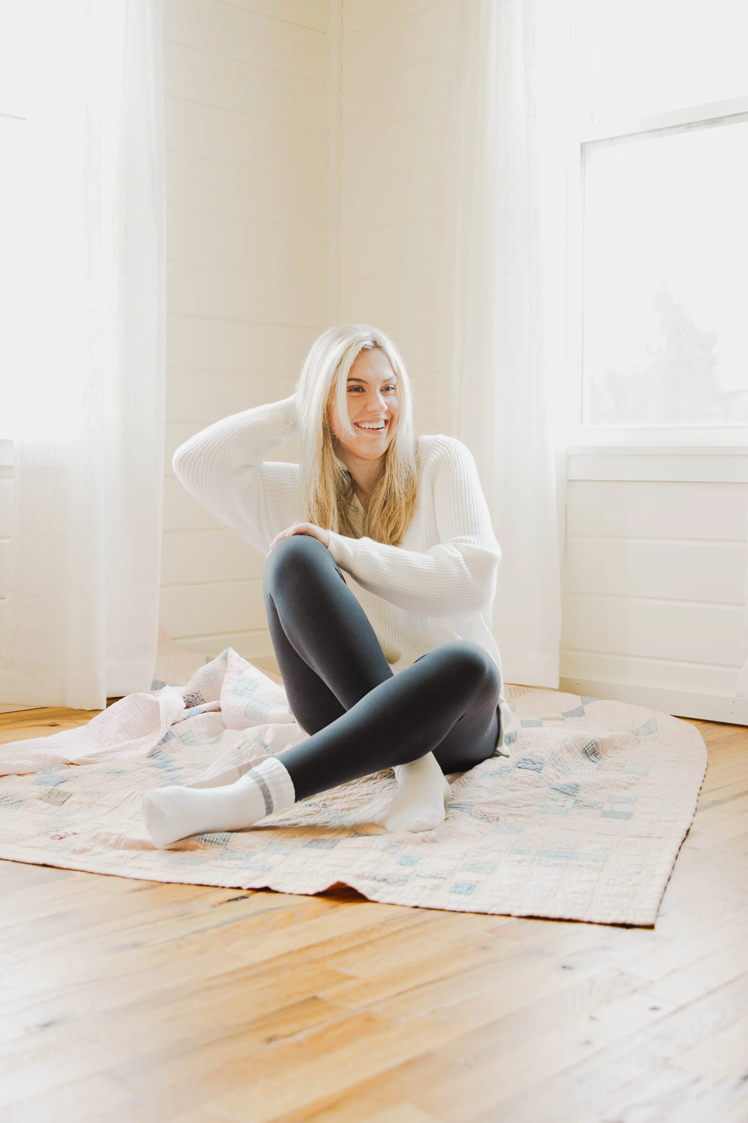 A young woman with blonde hair sitting on a quilted rug in a bright room with white walls and large windows, smiling and wearing a white sweater, black leggings, and white socks.