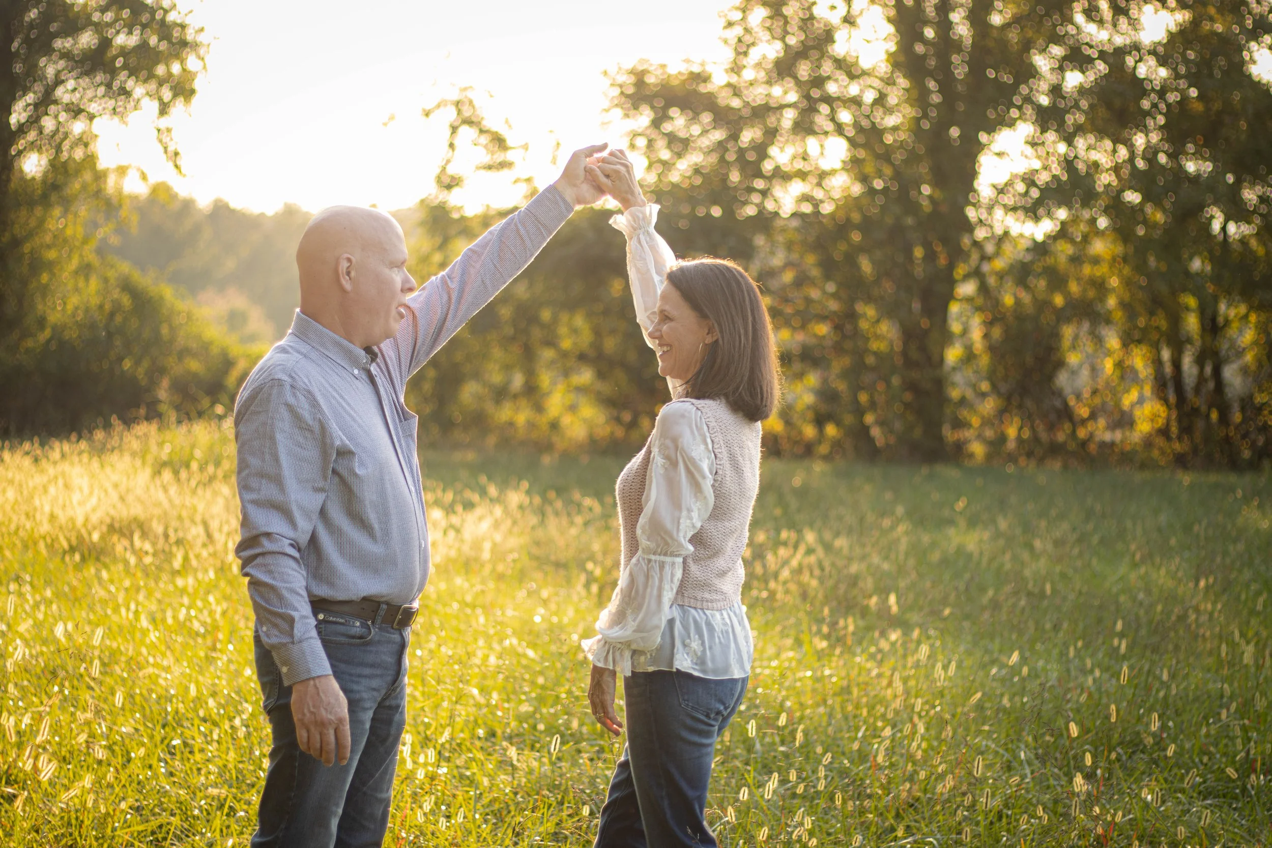 An older man and a woman are dancing in a sunlit field with trees in the background. The man is holding the woman's hand up as they dance, both smiling and enjoying the moment during sunset.