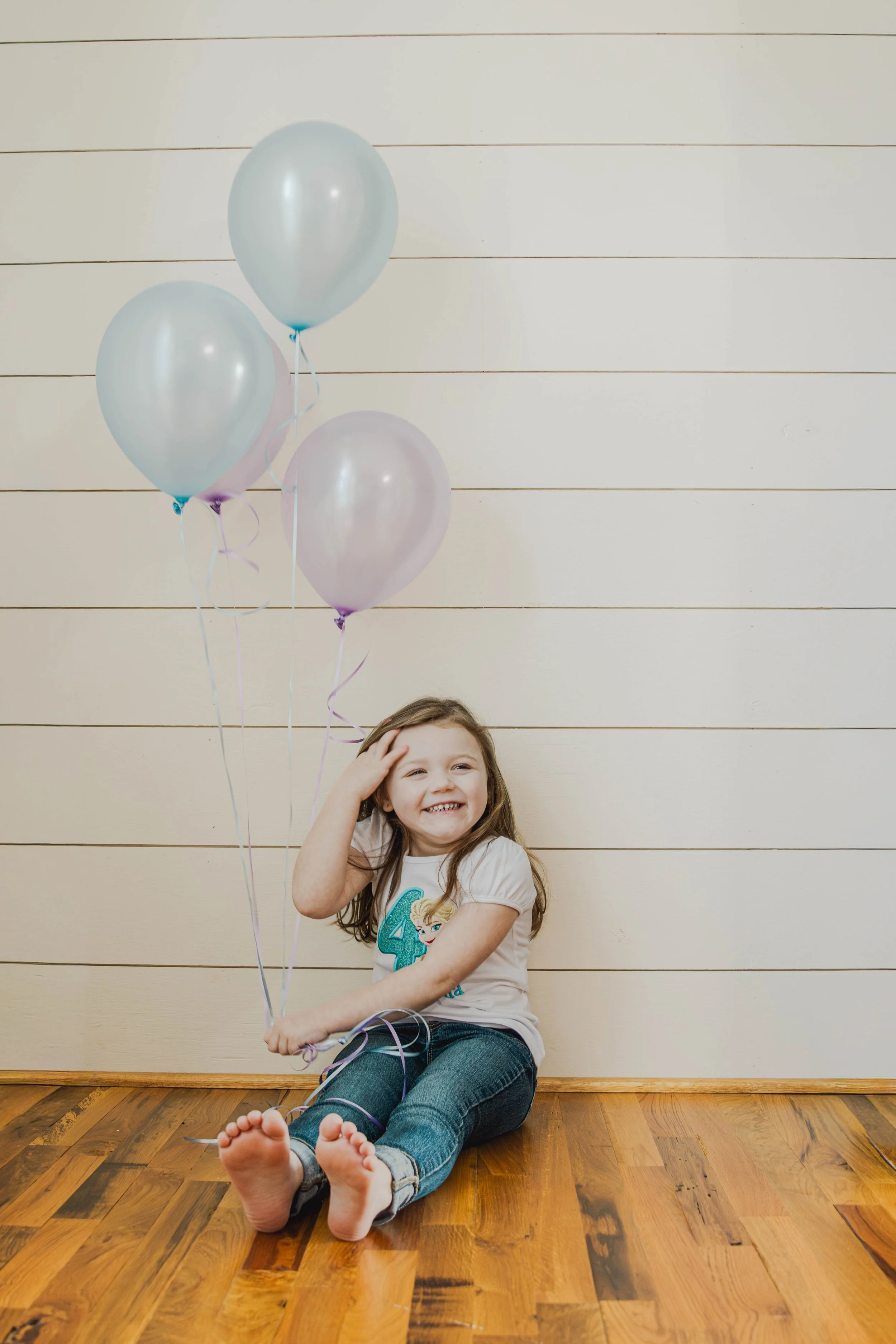 A young girl sitting on a wooden floor, smiling, with three balloons behind her. The balloons are pastel shades of blue and purple. The wall behind her is decorated with horizontal white wooden planks.