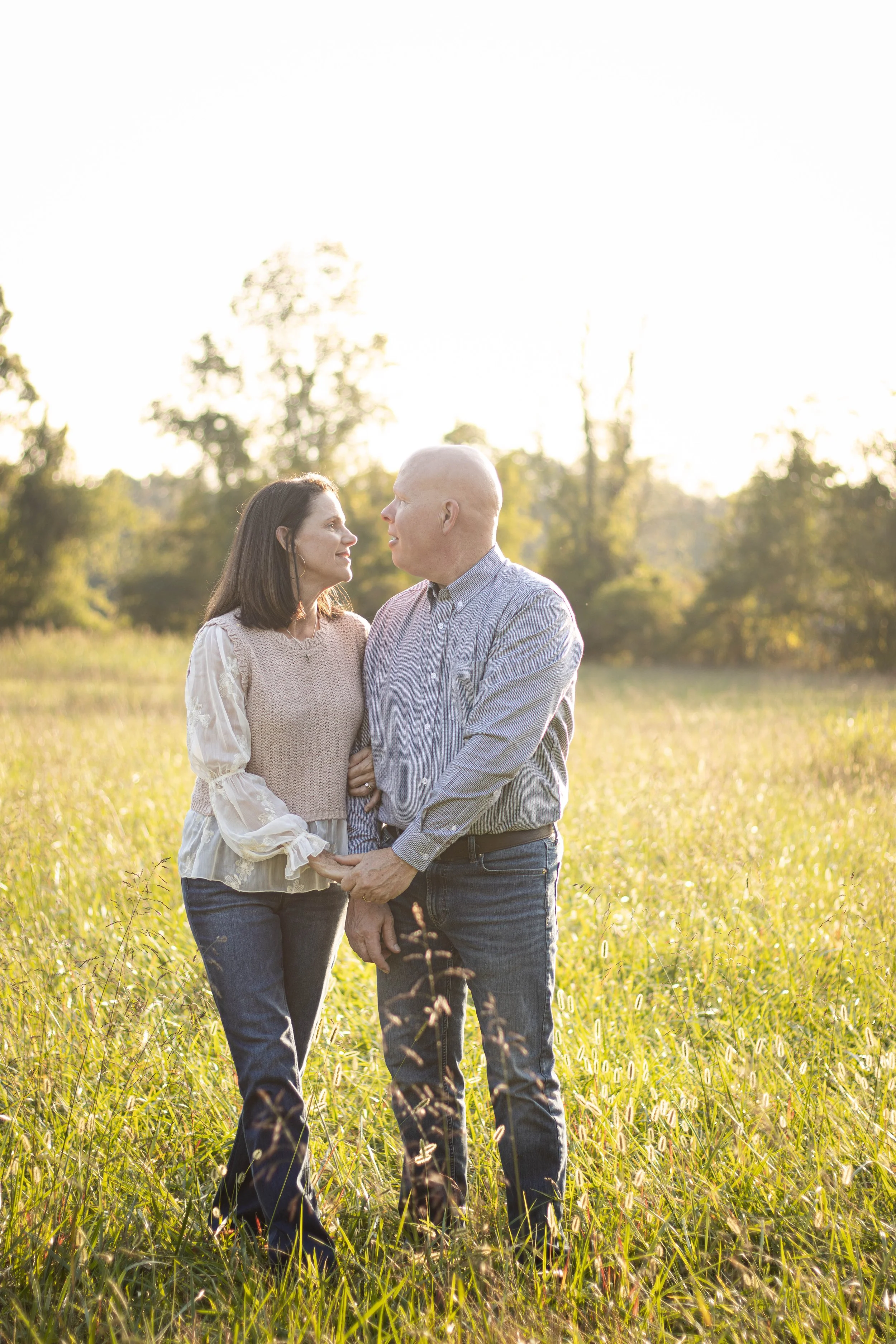 A couple holding hands and looking at each other in a grassy field with trees in the background during sunset.