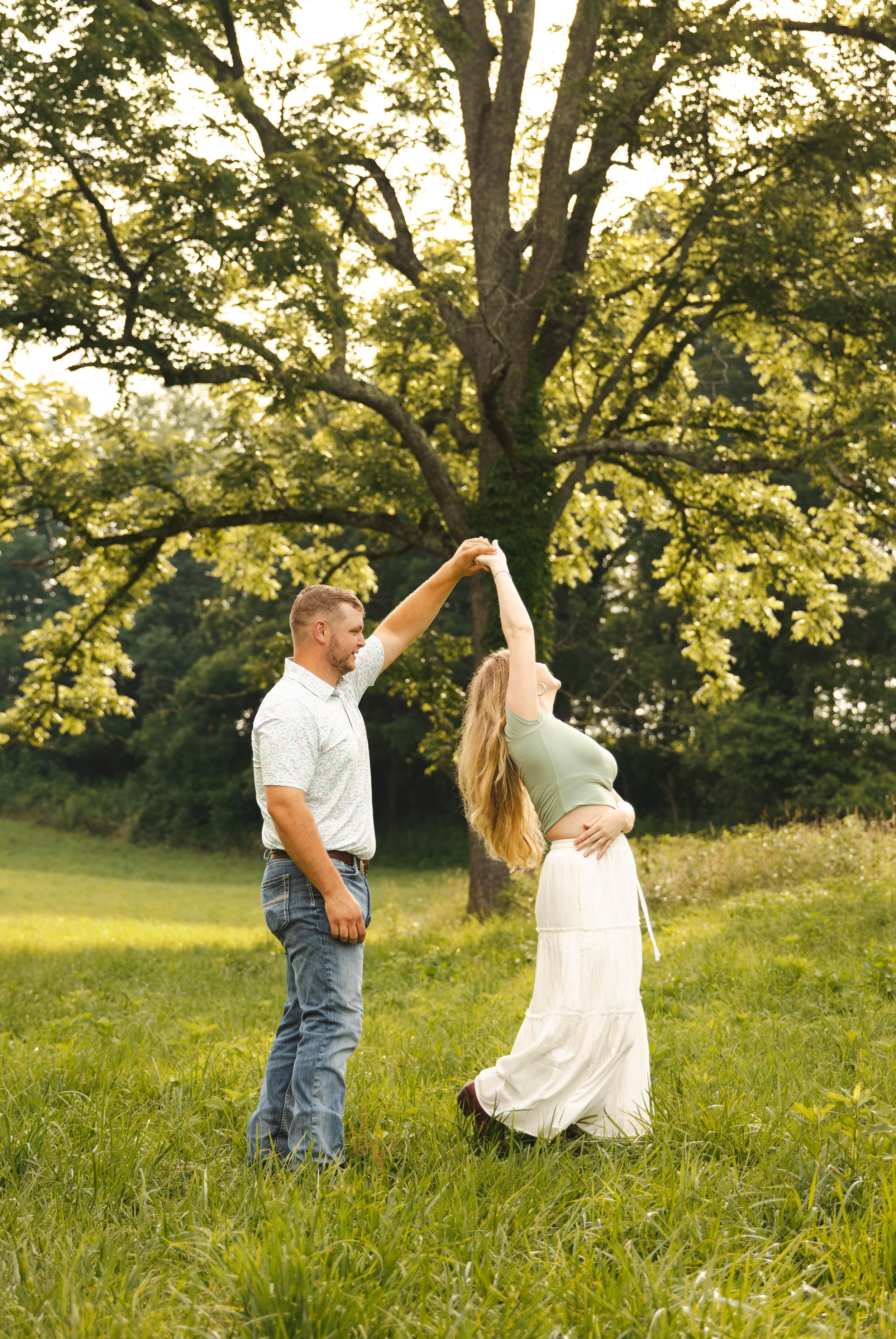 A couple dancing in a grassy field under a large leafy tree during daylight.