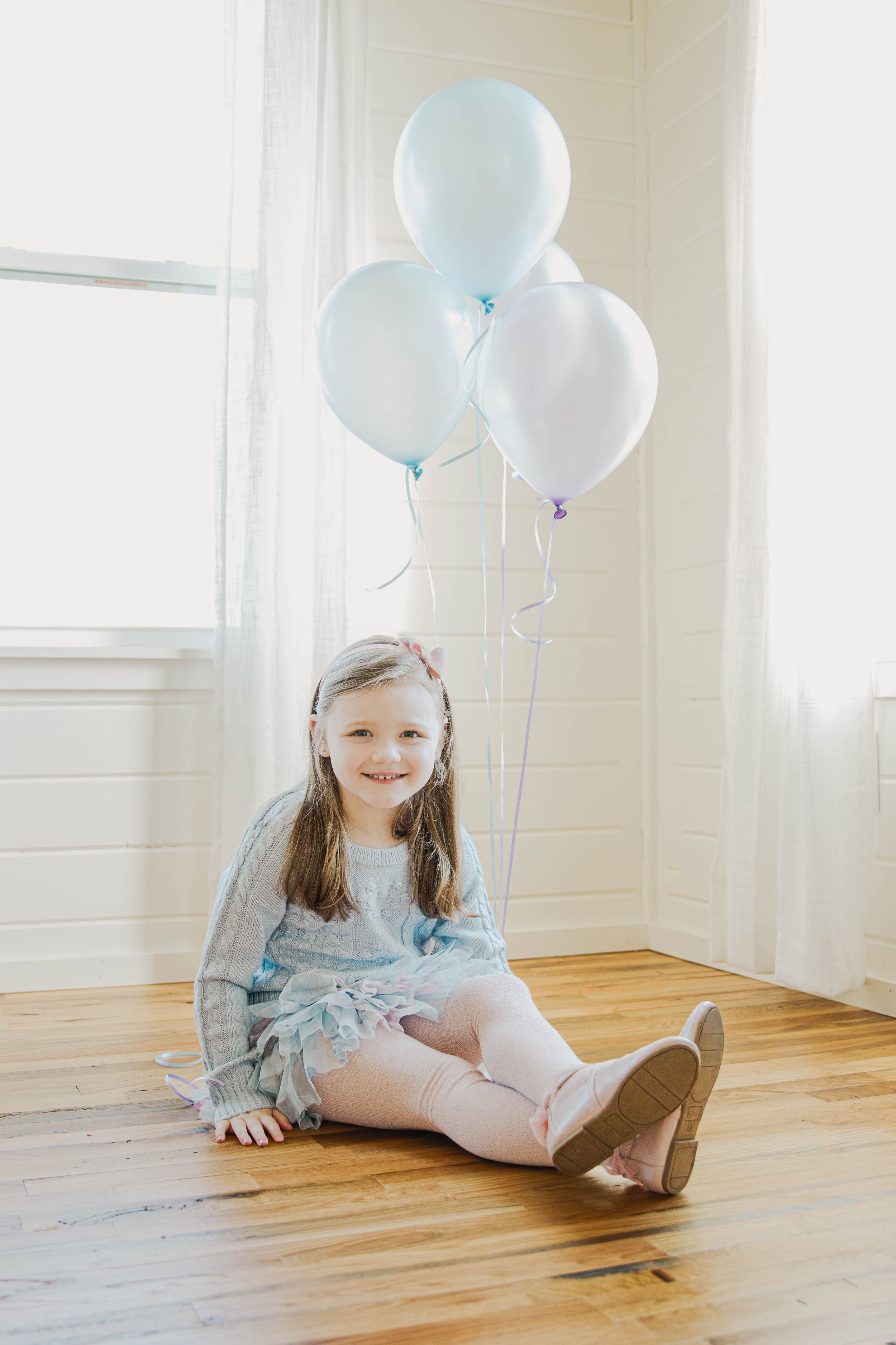 A young girl sitting on a wooden floor holding a bunch of light blue balloons. She is smiling and wearing a pastel-colored dress with tights. The room has white walls, curtains, and natural lighting.