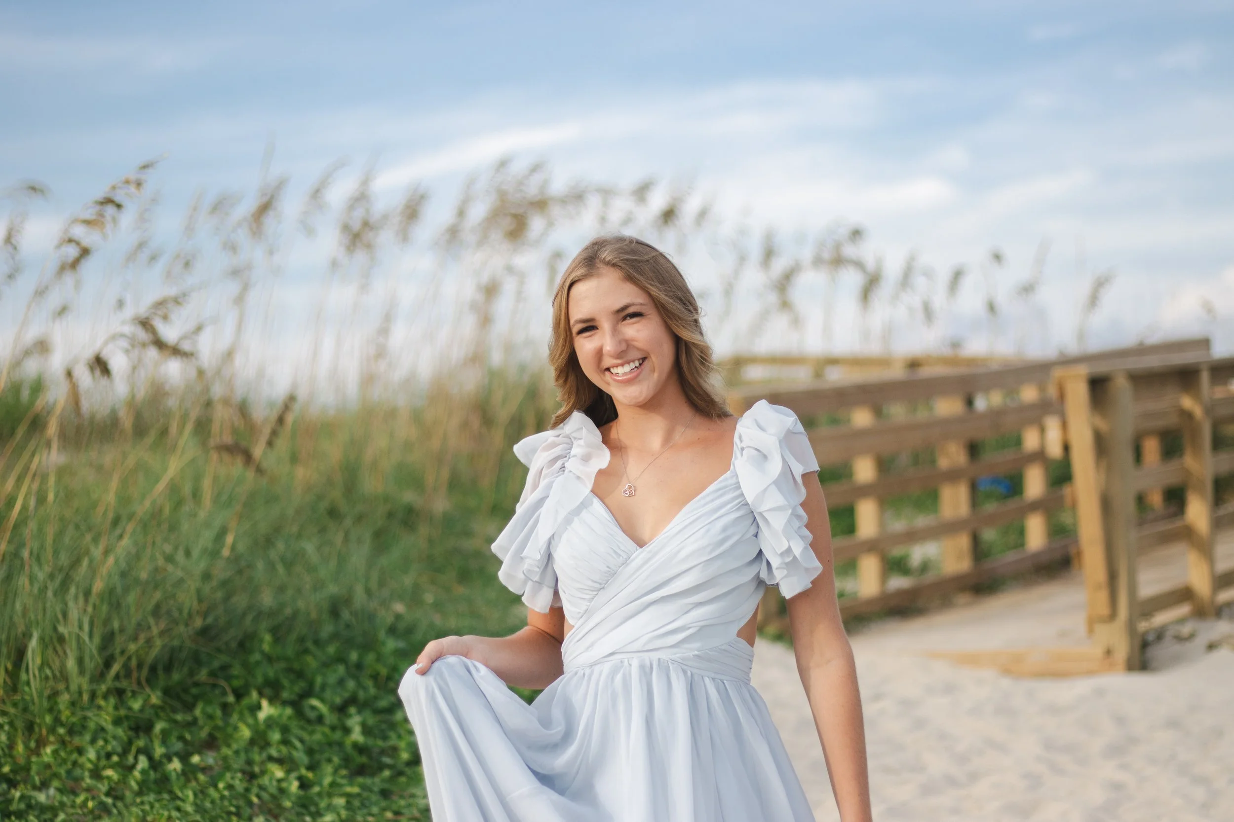 A young woman in a white dress smiling and standing outdoors on a sandy path with wooden railing and tall grass in the background.