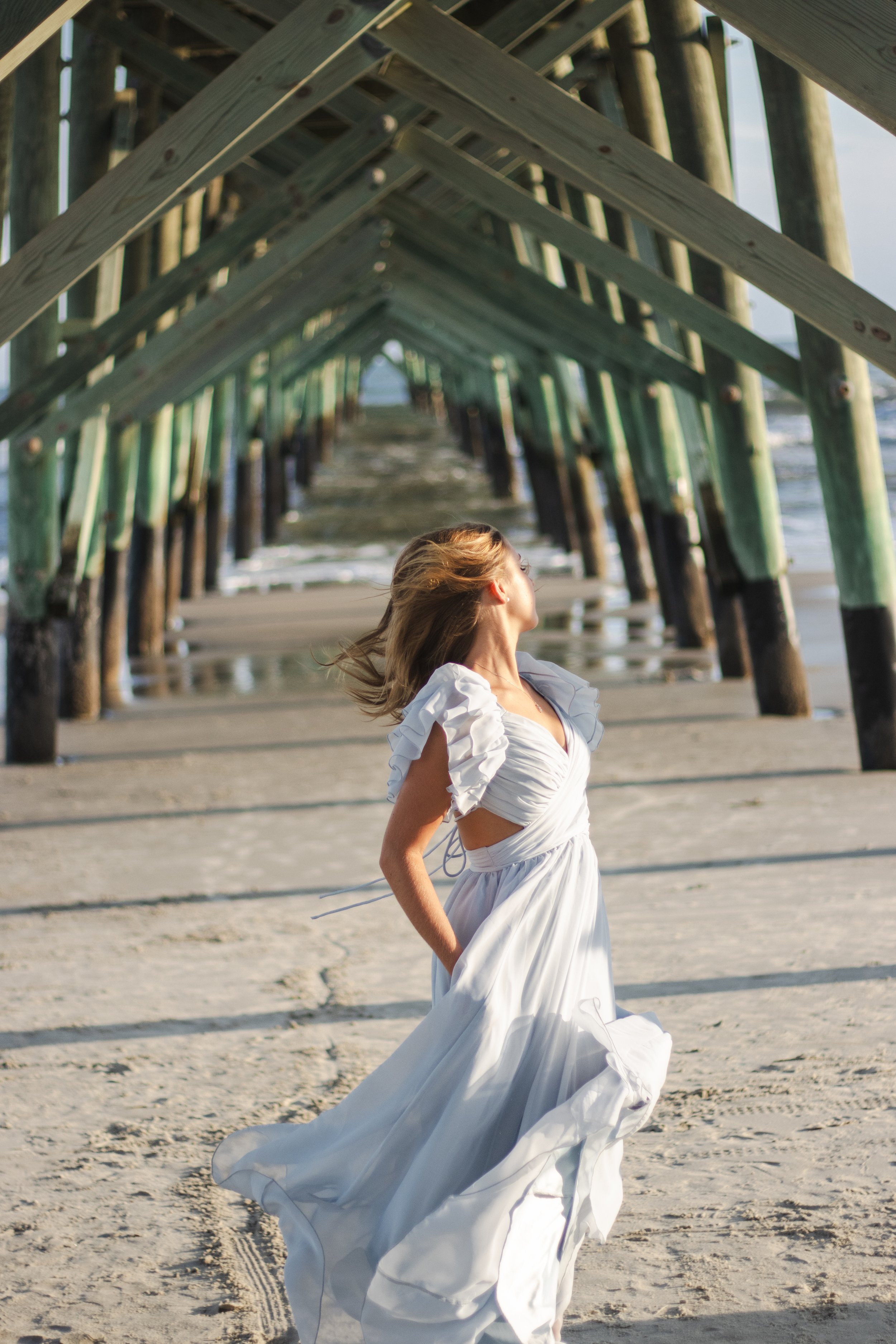 A woman in a white dress standing on a sandy beach under a wooden pier.