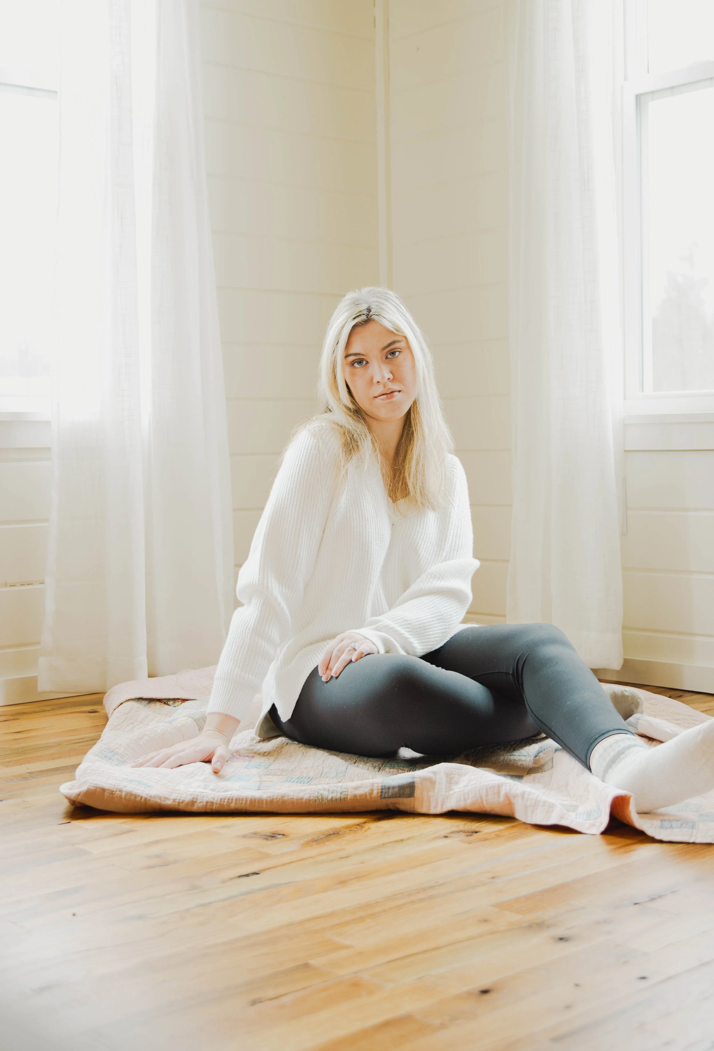 A young woman with blonde hair sitting on a quilted blanket on a wooden floor near large windows with white curtains, wearing a white sweater and black leggings.