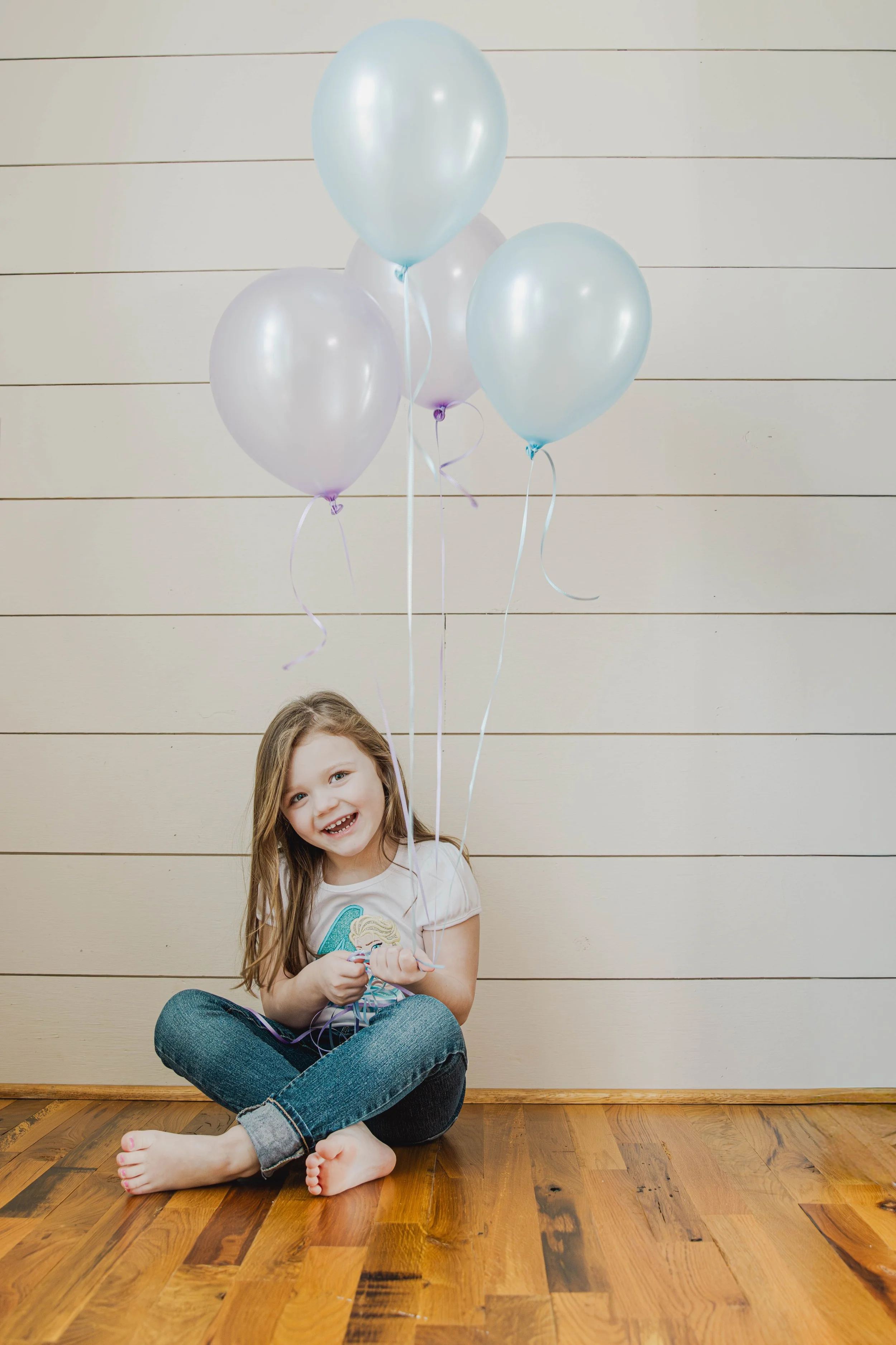 A young girl sitting on a wooden floor holding pastel-colored balloons in front of a light-colored paneled wall.