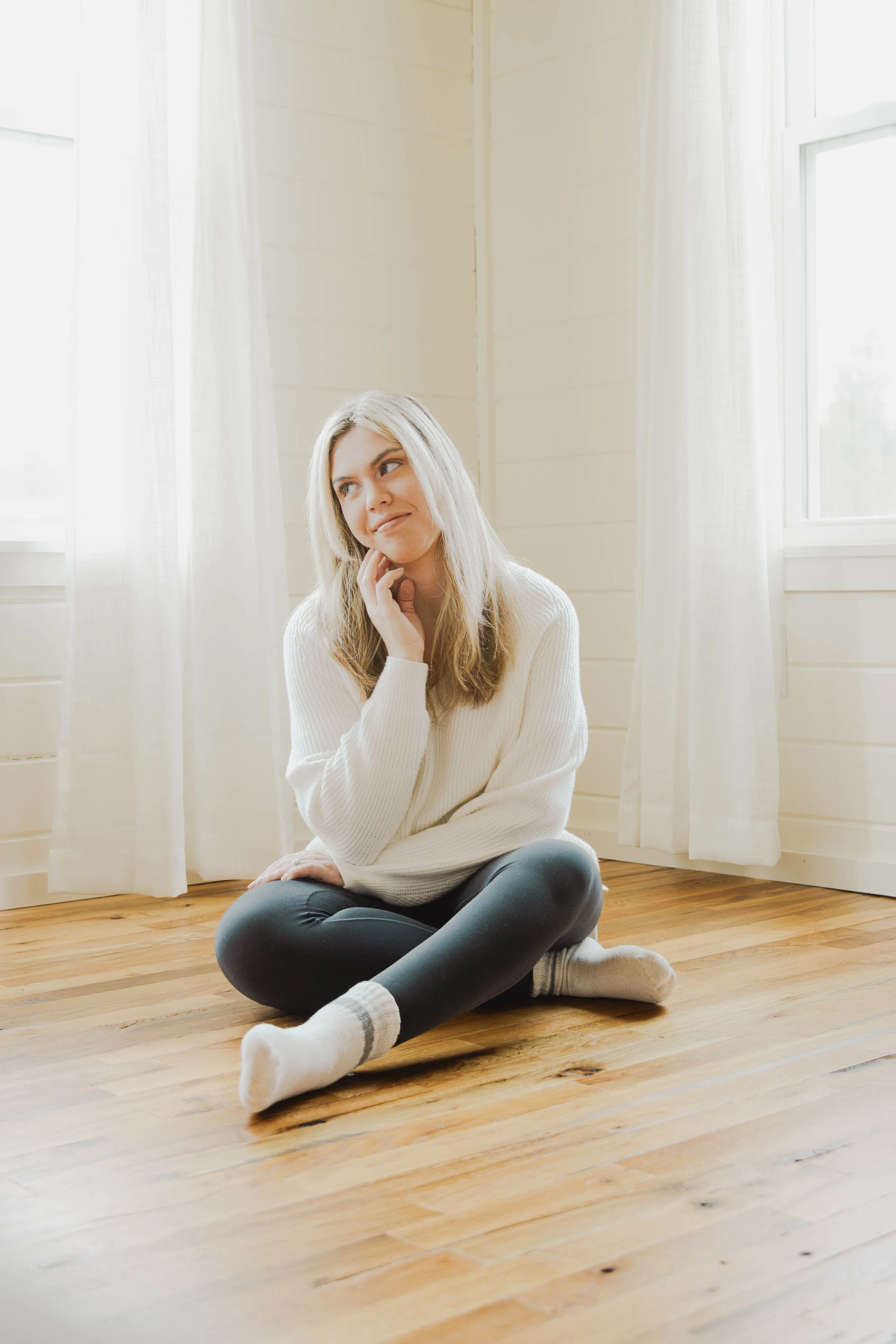 A woman sitting on a wooden floor, wearing a white sweater, dark pants, and socks, with her hand on her chin, looking thoughtfully to the side near white curtains and windows letting in natural light.
