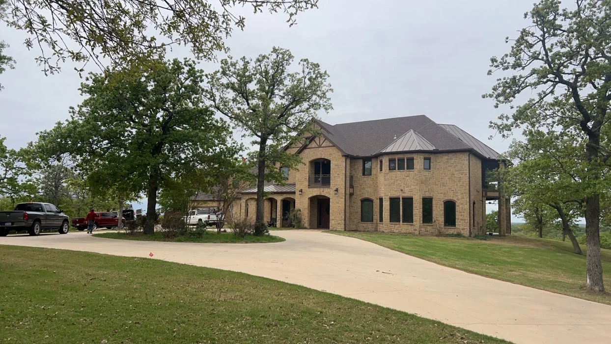 Large brick house with multiple levels and a sloped roof, surrounded by trees and a curved driveway.