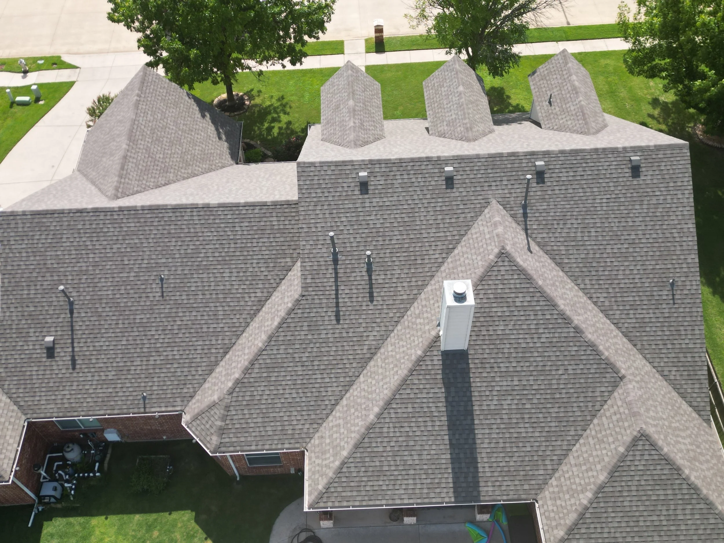 Aerial view of a house with multiple gable roofs, vents, and a chimney, surrounded by a green lawn and trees.