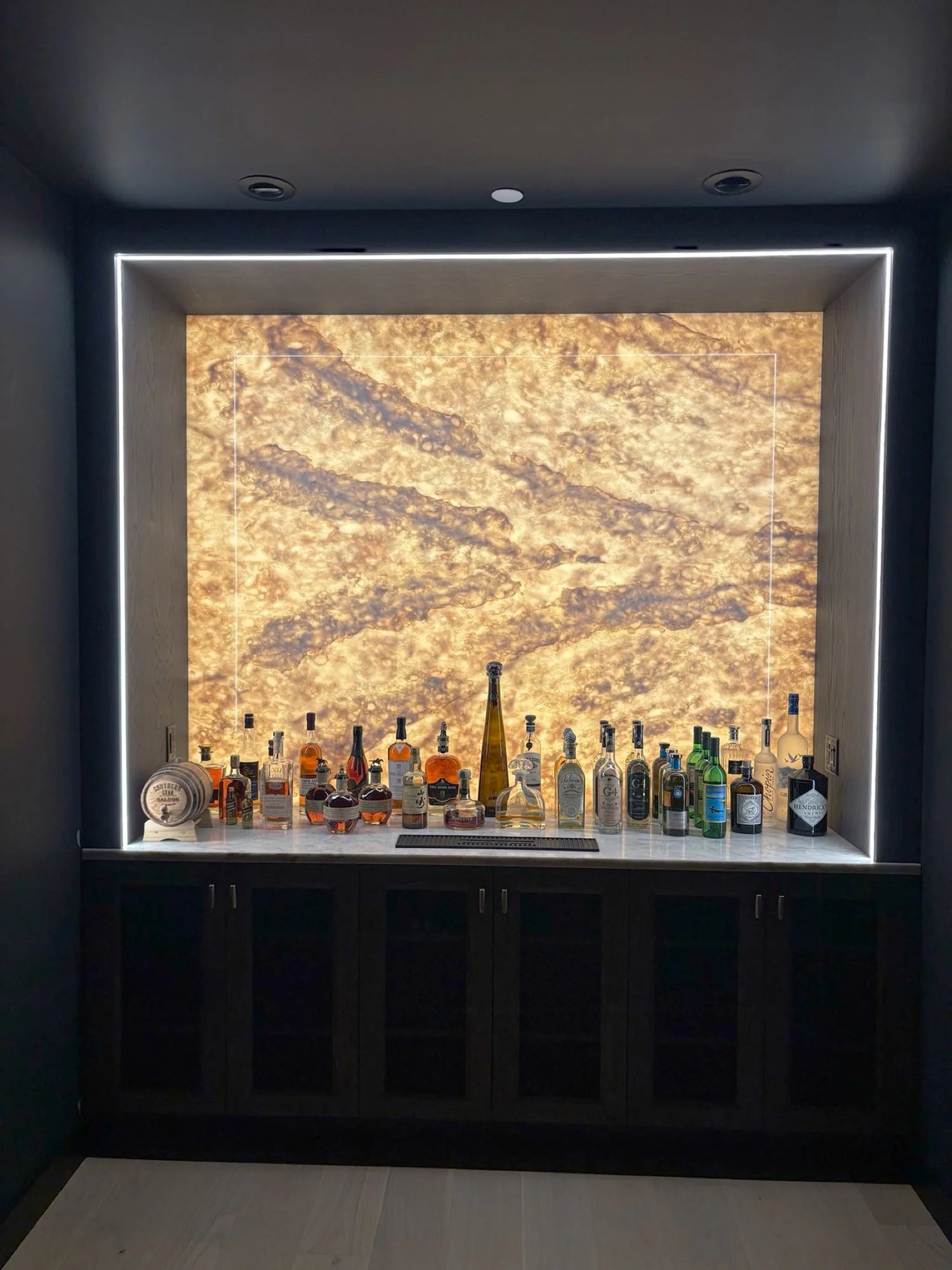 A bar with a backlit, marbled wall featuring various bottles of liquor and spirits on a white marble counter, below a dark ceiling with recessed lighting.
