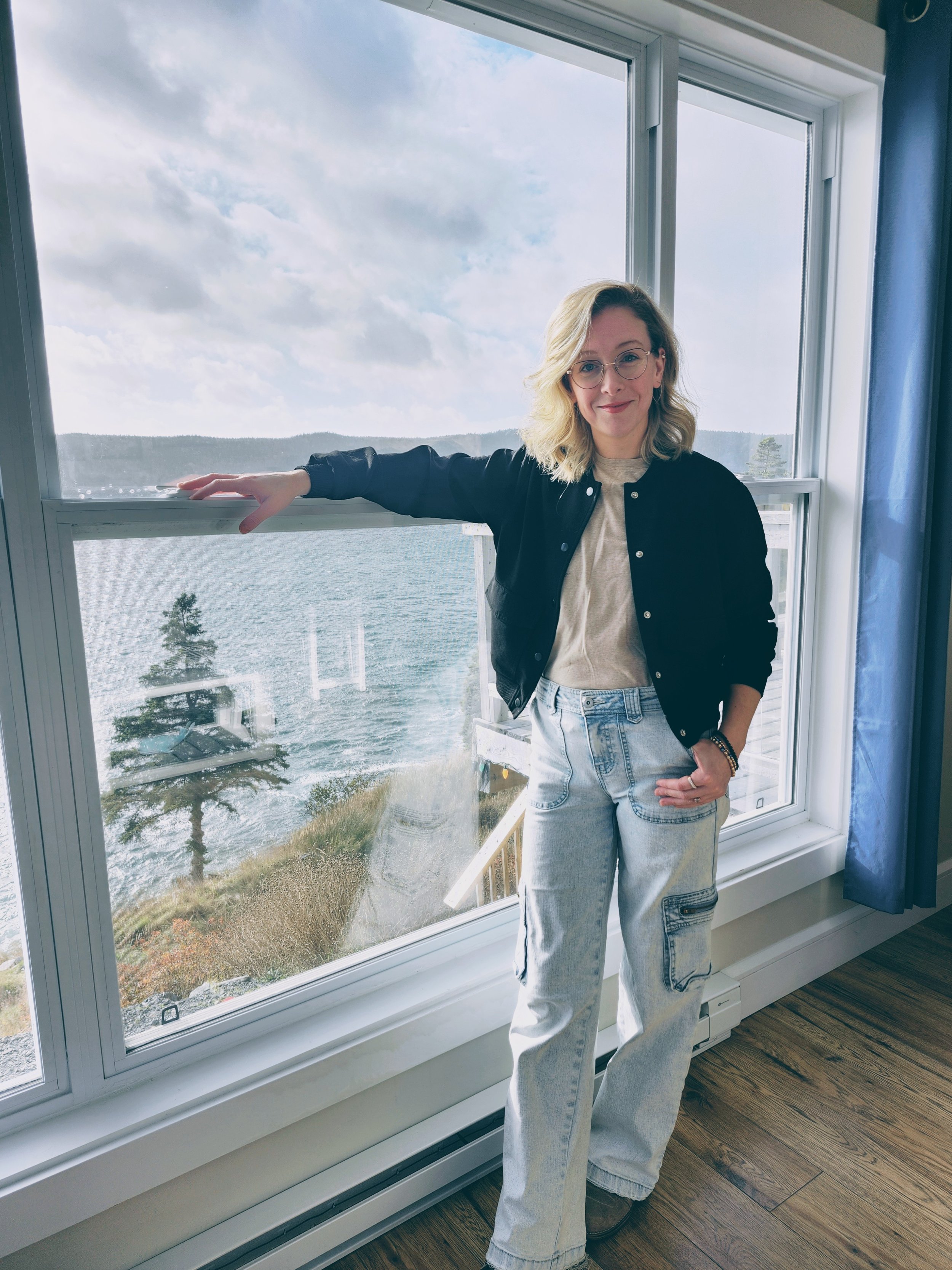 Woman standing indoors in front of a large window with a view of water and trees outside.