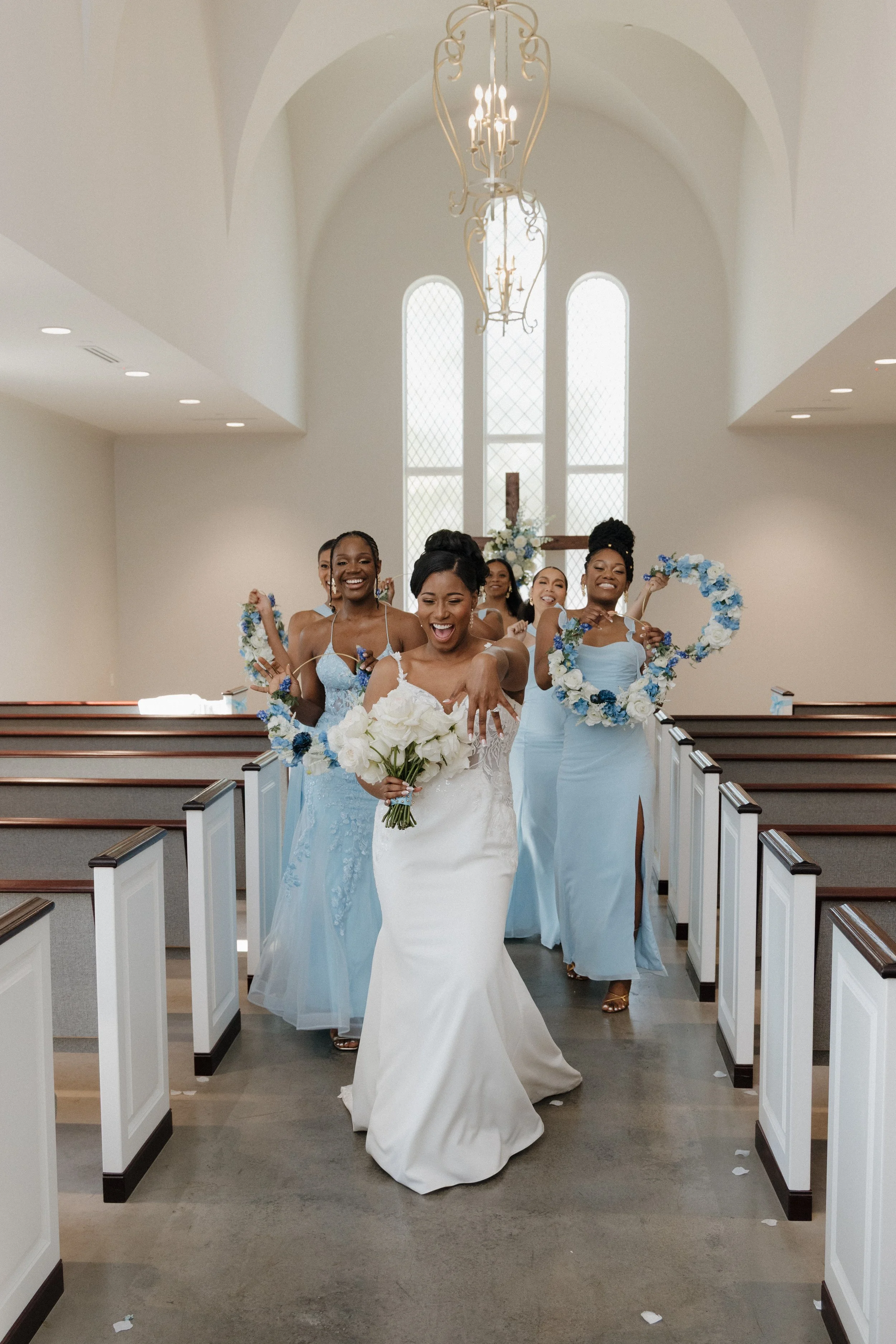 A bride and bridesmaids walking down the aisle of a church during a wedding ceremony, all smiling and holding floral bouquets, with a cross and large windows in the background.