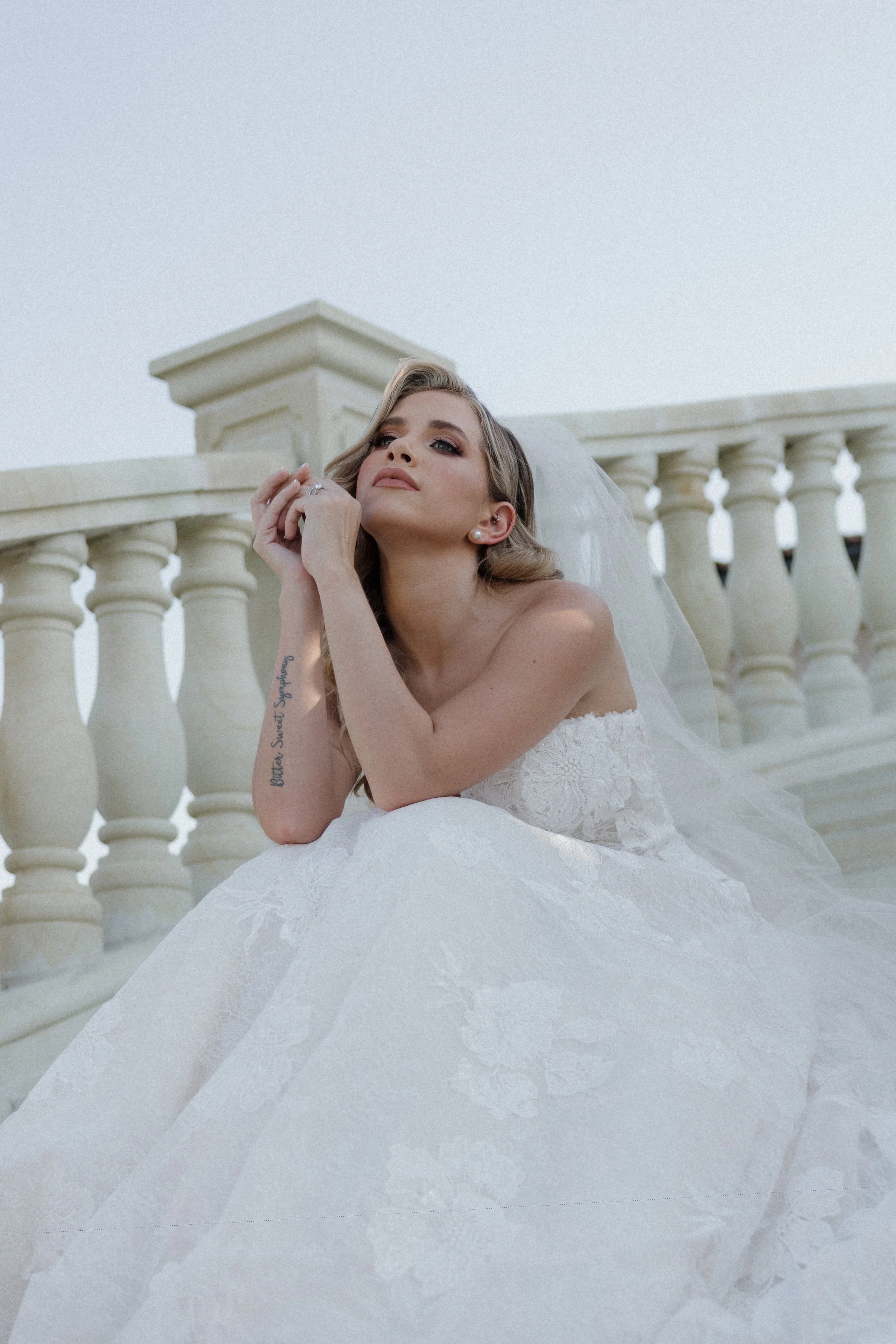 A woman in a white wedding dress sitting on a beige stone balcony with balusters, looking confidently at the camera with her hand near her face.
