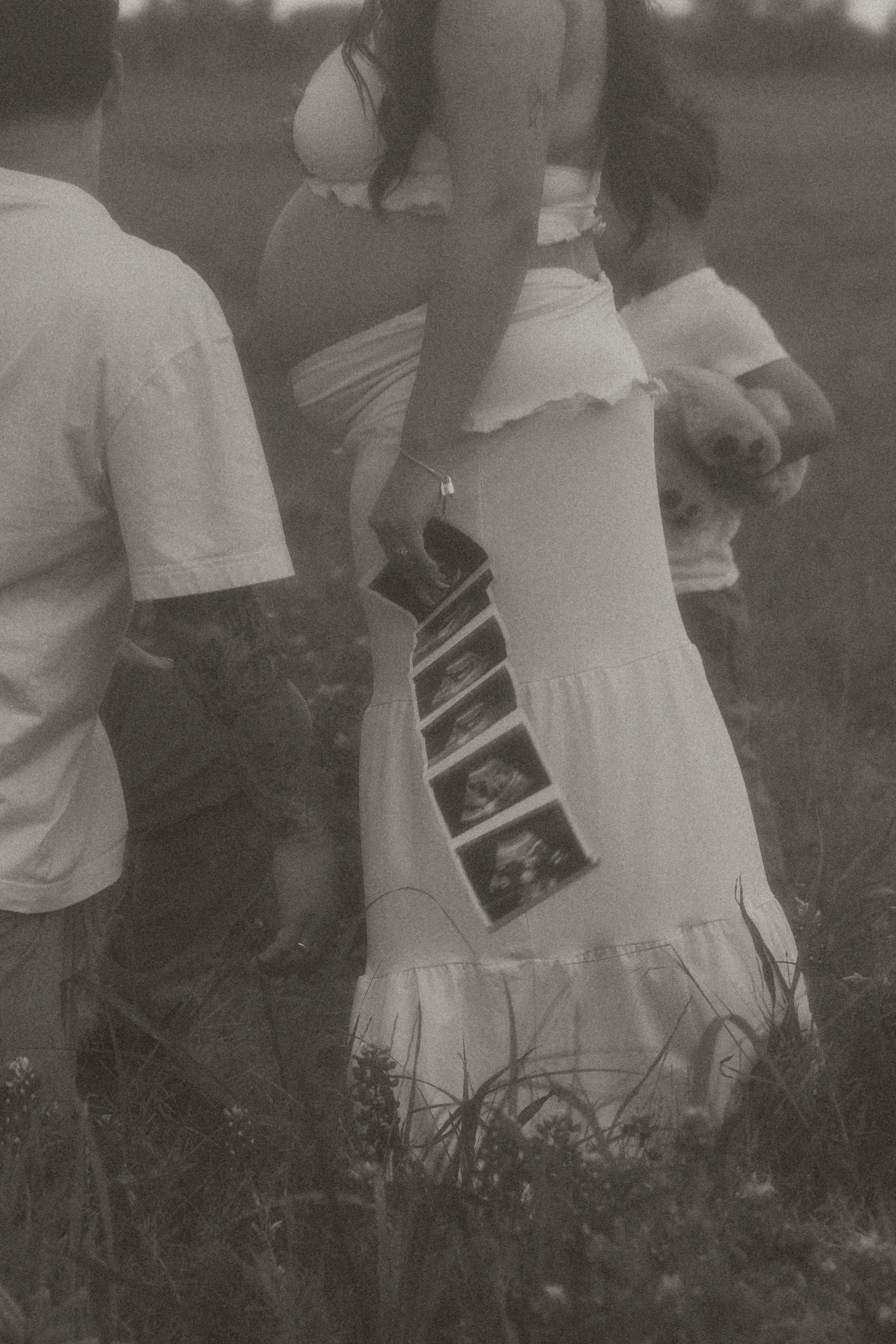 Black and white photograph of a person holding a photo album, with a dog lying on the grass nearby, and another person sitting on the grass in the background.