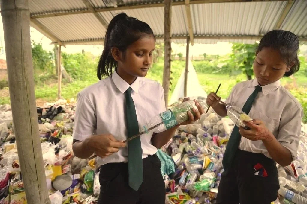 Two girls in school uniforms with white shirts and green ties are collecting plastic bottles in a trash yard, using sticks to pick up the bottles. They are standing under a makeshift shelter with a corrugated metal roof. The background shows a pile of scattered plastic bottles and some greenery.
