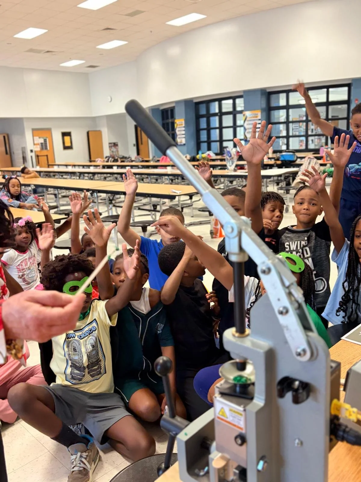 Group of children in a classroom or cafeteria raising their hands, with a microscope in the foreground, some children wearing green glasses, and a woman holding a stick or pointer.