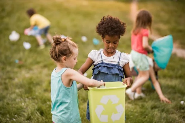 Three young girls outdoors, one holding a yellow recycling bin, others collecting trash, in a grassy area