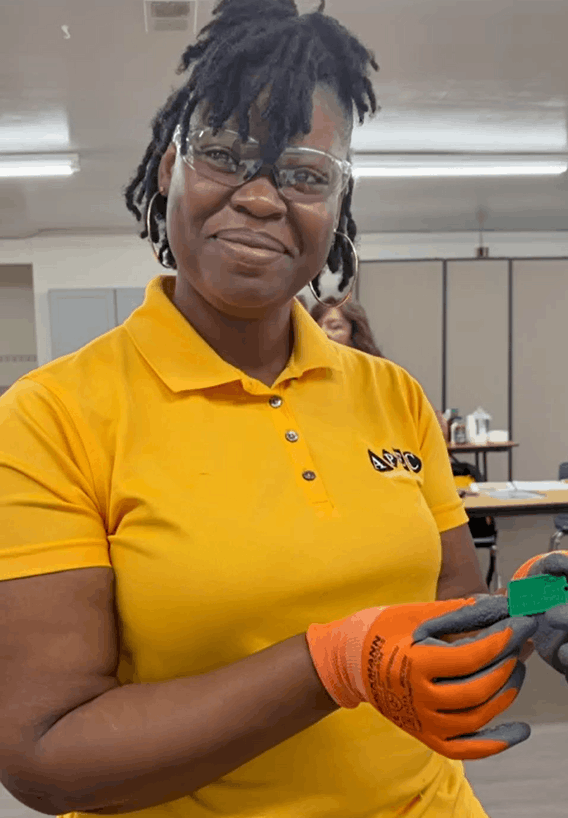 A woman in safety glasses and orange work gloves holding a small green object in a classroom or workshop setting.