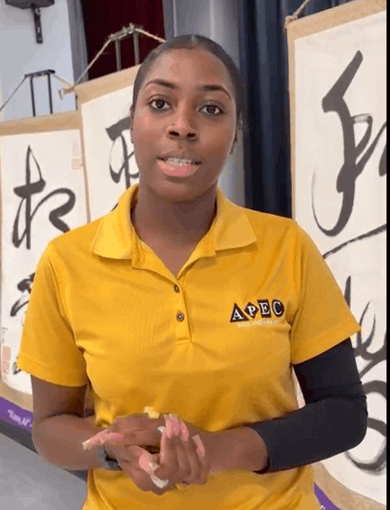 Young woman in a yellow polo shirt with APEC logo, standing indoors in front of banners with Asian characters, holding her hands together with some pink paint on her fingers.