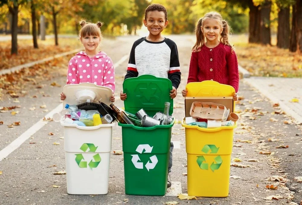 Three children stand outdoors on a fallen autumn leaves path, each holding a recycling bin filled with recyclable household items.