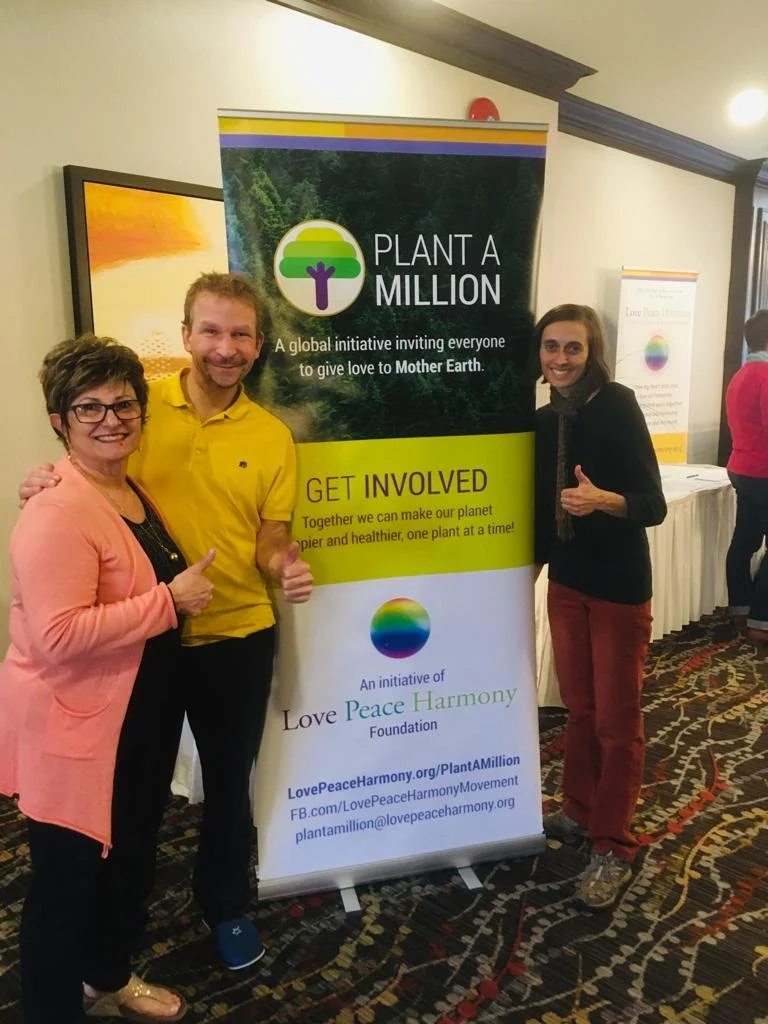 Three people standing next to a banner promoting a global environmental initiative called Plant A Million, with the slogan 'A global initiative inviting everyone to give love to Mother Earth.' The banner encourages involvement to make the planet healthier, and mentions the Love Peace Harmony Foundation.