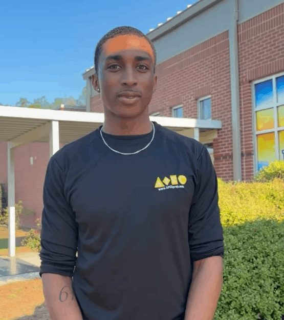 Young man standing outdoors in front of a brick building, wearing a black T-shirt with yellow logo, silver chain, and short hair.