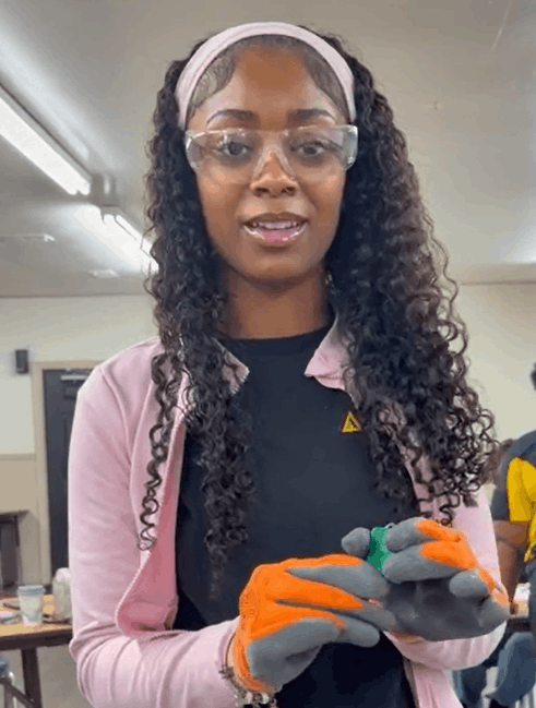 Young woman wearing safety goggles, gloves, and a pink jacket, holding a small object in her hands, with a classroom or laboratory setting in the background.