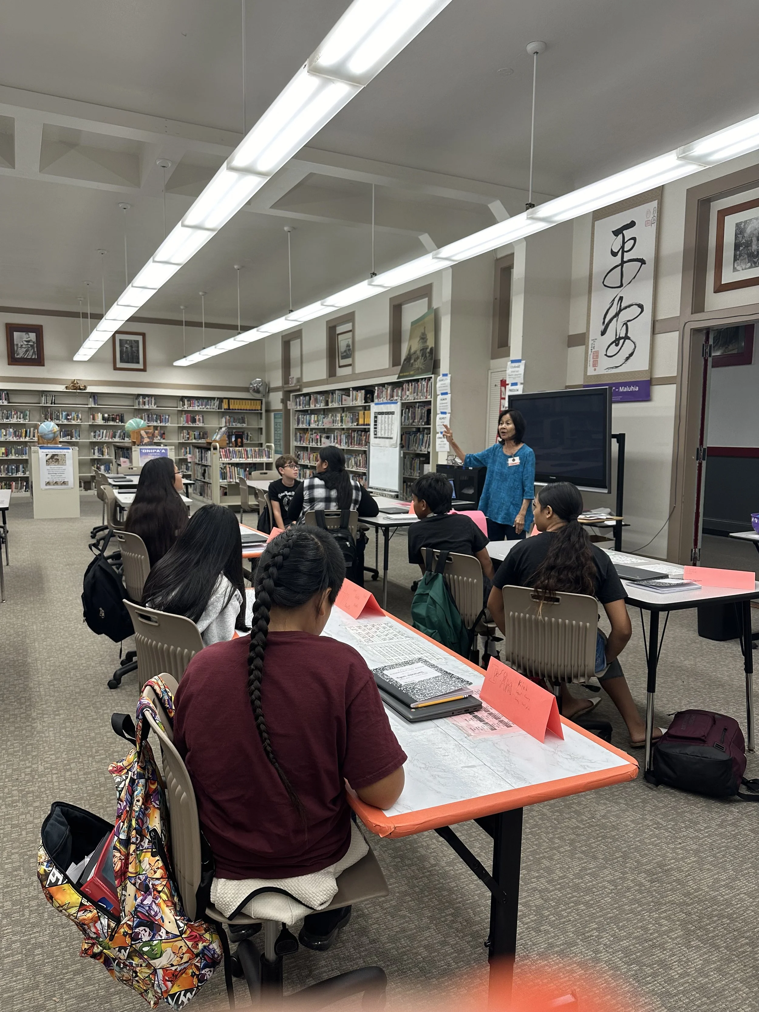 A classroom or library setting with a teacher at the front and students seated at tables, listening to the teacher.