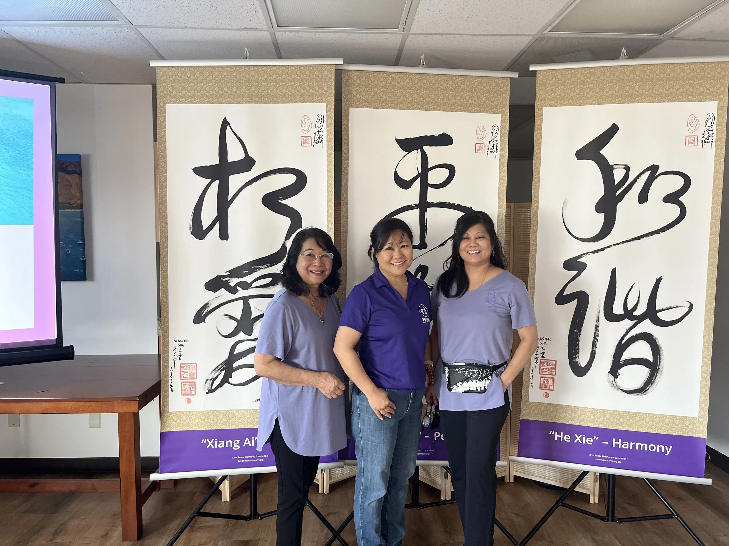 Three women standing together in front of large calligraphy banners with Chinese characters, smiling at the camera.